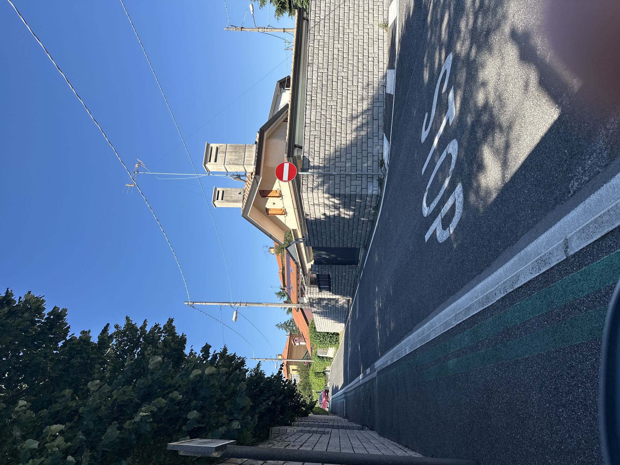 The image captures a quiet, tree-lined residential street in Trieste, Italy, on a clear, sunny day. The road, paved with asphalt, features white lane markings and a distinctive triple green line on the left side, likely indicating a parking or no-stopping zone. In the foreground, the word "STOP" is stenciled in white capital letters on the road surface, casting dappled shadows from overhead foliage.
A red car is visible further down the street, parked or slowly moving. The street is bordered by high, grey stone walls on both sides, with a row of houses with terracotta tiled roofs visible behind the wall on the right. One house has twin chimneys and a satellite dish mounted on its roof. A circular traffic sign with a red border and a white horizontal bar, indicating "No Entry" or "Do Not Enter," is positioned on a pole near the corner of the stone wall.
A large tree with dense green foliage partially shades the left side of the street and the stone wall. Overhead, power lines and a streetlamp are visible against the bright blue sky. The scene suggests a peaceful, suburban environment within Trieste.