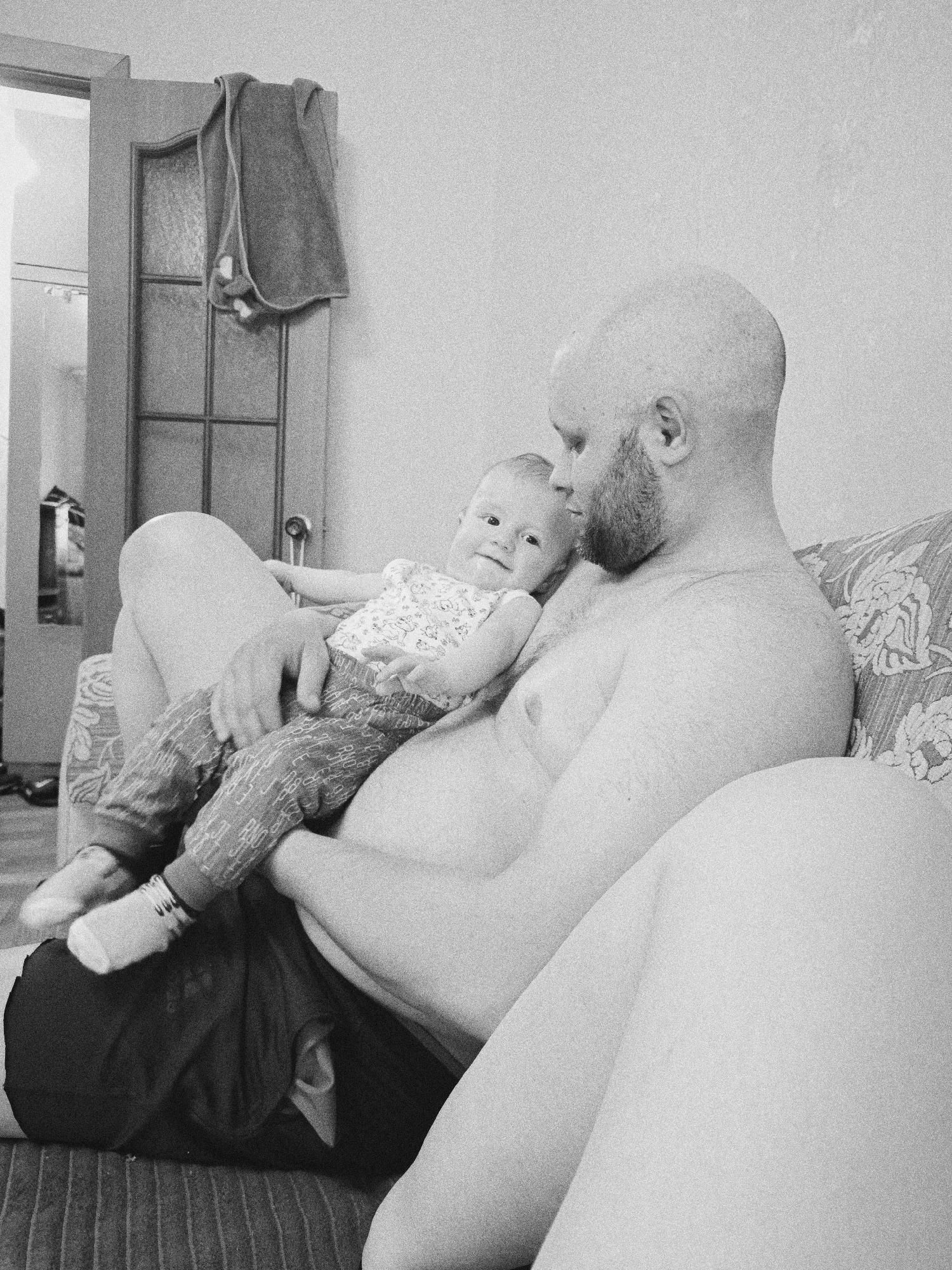 A shirtless, bald man with a beard sits on a couch and holds a baby in his lap. The baby is wearing patterned pants and socks. A door with a towel hanging on it is visible in the background. The scene is in Tobolsk, Russia.