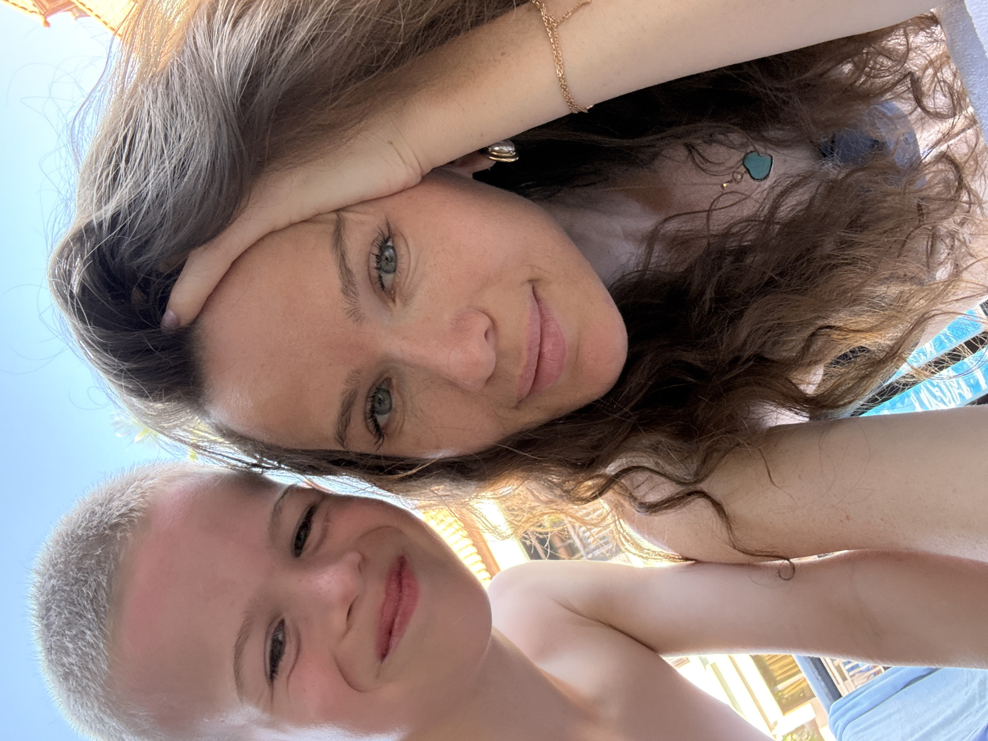 A selfie is taken outdoors, with a clear blue sky above. On the left side of the frame is a young boy with short, light-colored hair, looking directly at the camera and smiling. He is shirtless. On the right side of the frame is a woman with long, wavy brown hair, her right hand touching her forehead. She also looks at the camera, with a slight smile. She is wearing a light-colored top and a necklace with a turquoise pendant. Her left arm is visible, with a thin bracelet on her wrist. The background shows a blurry view of buildings with balconies and some greenery, suggestive of a residential or hotel area. The lighting suggests it is daytime, possibly late afternoon due to the warm tones. The overall impression is a casual, relaxed moment. This scene is not identifiable as being located in Dubai, United Arab Emirates.