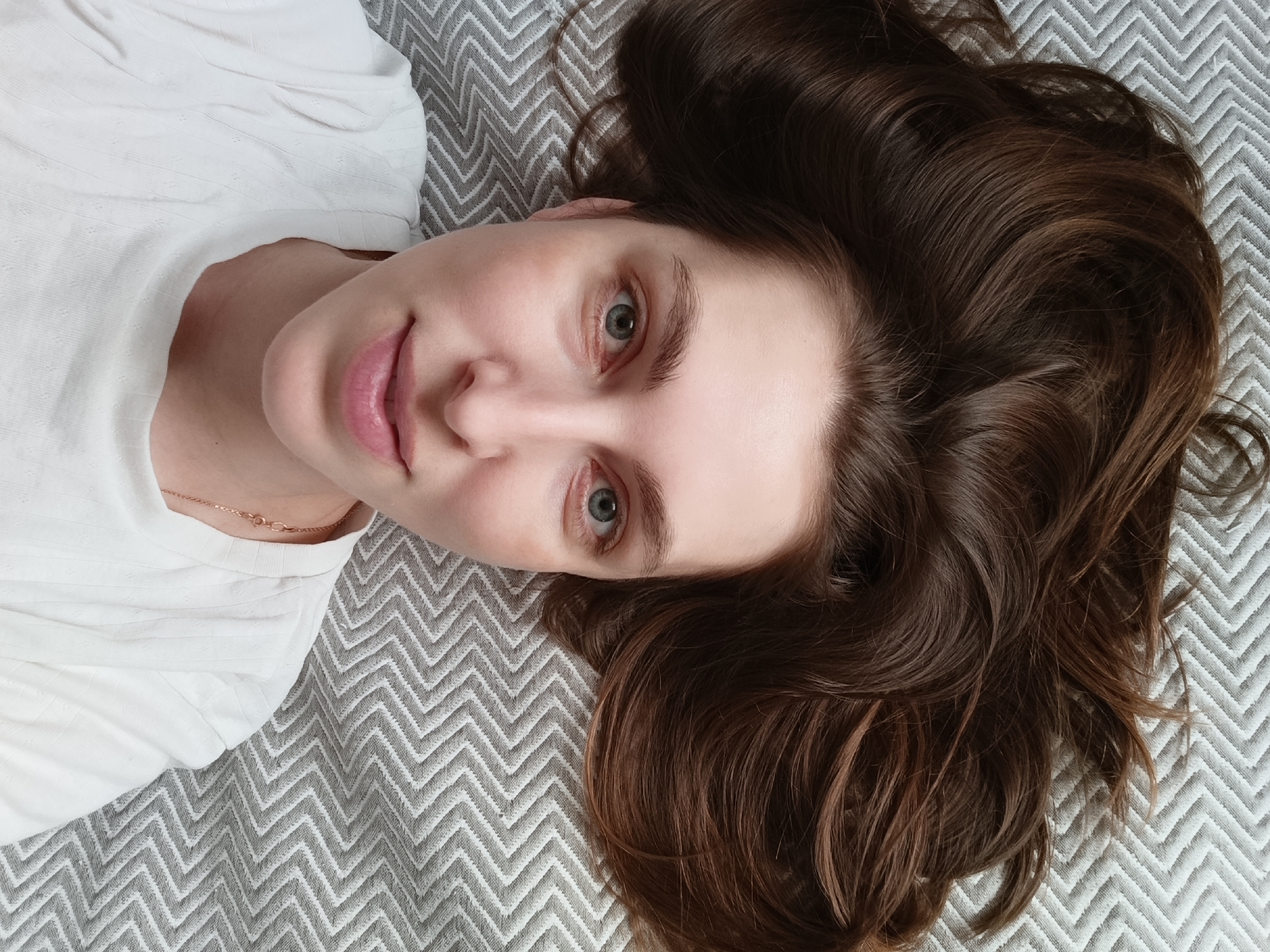 A young woman with long, auburn hair is lying on a gray and white zigzag-patterned surface. She is wearing a white shirt and a delicate gold necklace. She is looking directly at the camera with a slight smile. Her eyes are light blue. The photo appears to have been taken from a high angle. There is no immediate indication of the image's location being in Chaikovsky, Russia; there are no identifying features.