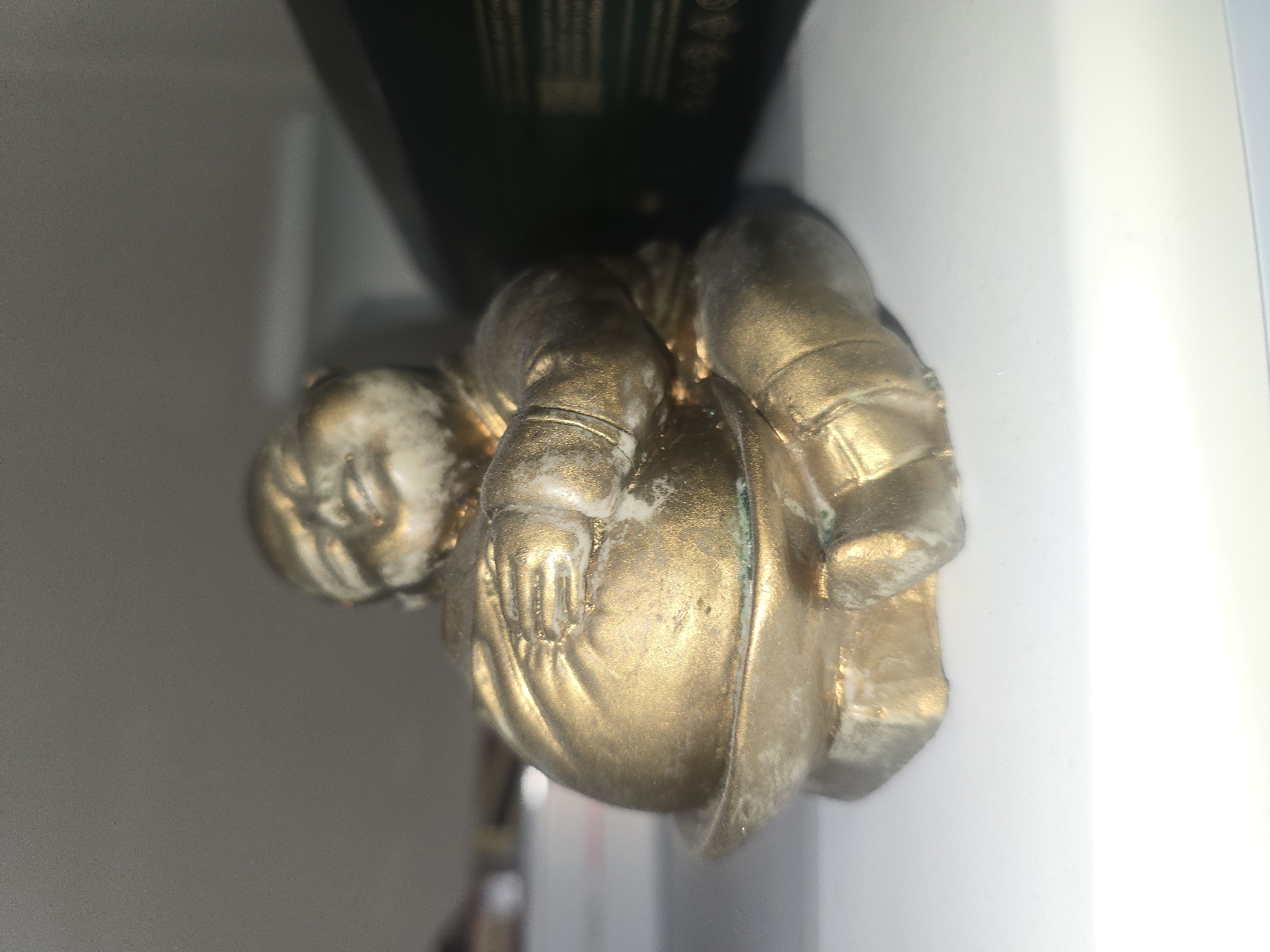 A small, golden, seated figurine is depicted. The figurine has a rounded belly and appears to be made of metal, possibly brass or bronze, with some areas showing tarnishing or discoloration. It is positioned on a white surface, likely a shelf or table. Behind the figurine, a dark object, possibly a book or box, is partially visible. The background is a muted gray, suggesting an indoor setting. The lighting is soft, casting a slight shadow to the right of the figurine. There is no information in the image to suggest a location in Ulan-Ude, Russia.