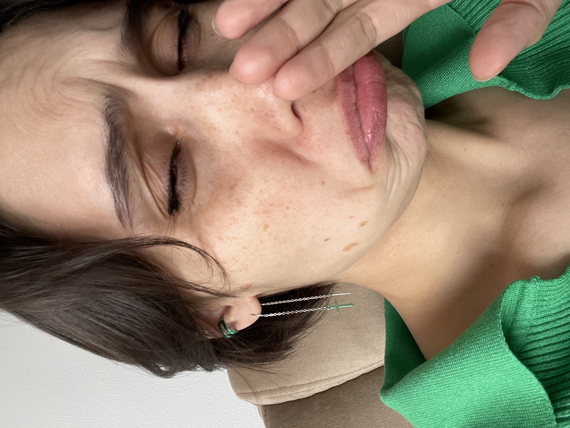 A close-up, rotated portrait captures an individual, likely female, from the chest up, in an indoor setting in Izhevsk, Russia. The person's eyes are closed, and their right hand is gently placed over their nose and mouth, suggesting a moment of introspection or shyness. They wear a vibrant green ribbed knit top. Visible facial features include dark, short hair, prominent freckles across the nose and cheeks, and full, reddish-pink lips. The left ear is adorned with two distinct earrings: a small green hoop and a longer silver chain earring with a dangling green cross charm. The background is simple, featuring a light-colored upholstered surface, possibly a sofa or chair, and a plain light wall. The lighting is soft and even, highlighting the textures of the skin, hair, and clothing. No text is visible in the image.