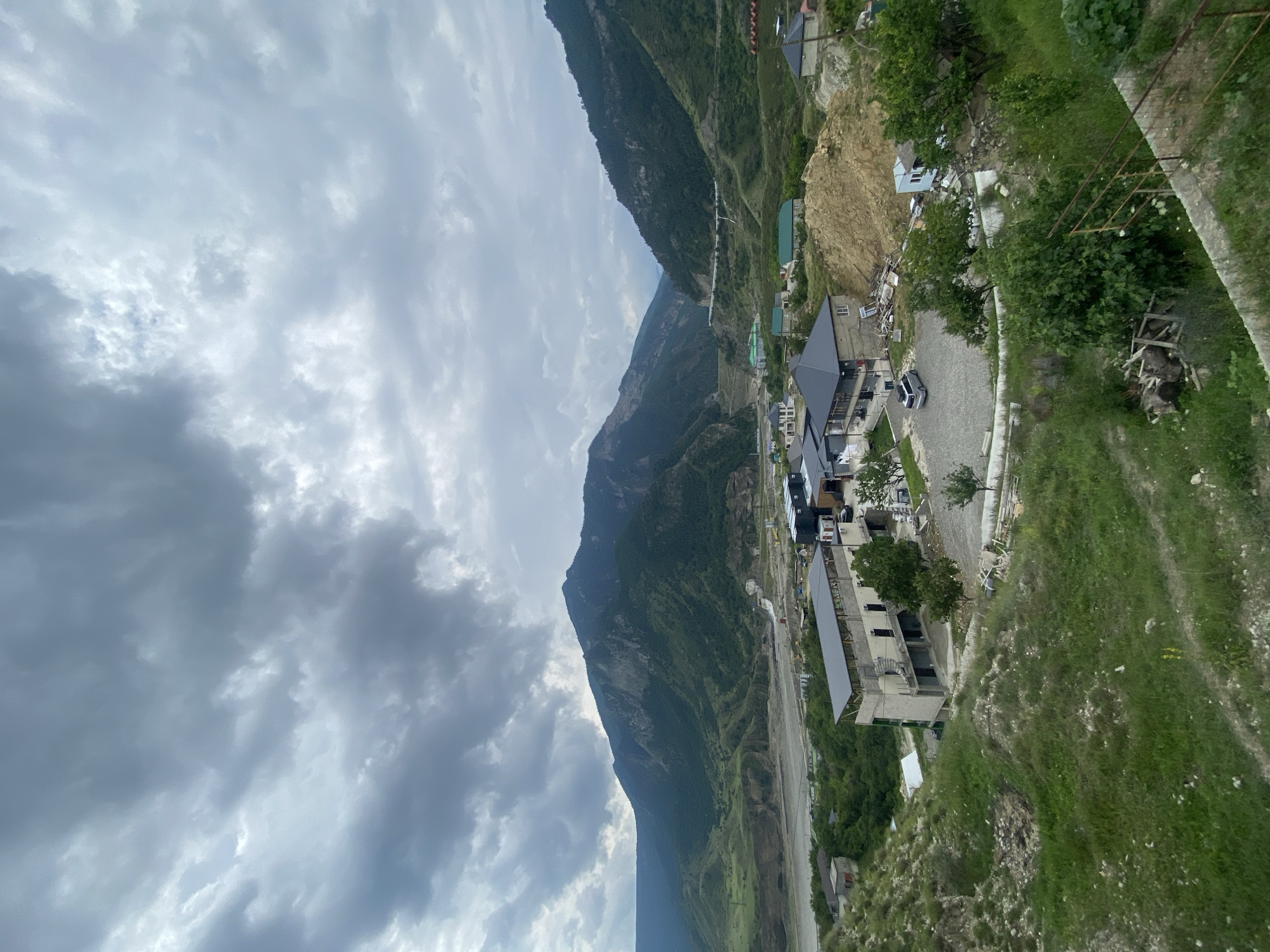 A wide-angle, high-angle shot captures a village nestled in a valley between large, verdant mountains under a cloudy sky. In the foreground, a grassy, uneven slope leads down to a collection of buildings. Several multi-story structures are under construction, with exposed concrete and scaffolding visible. One completed building has a dark, pitched roof and appears to be a residential or commercial property. A gravel parking lot next to it holds a single silver SUV. Further into the scene, more buildings with various roof colors are visible, along with trees and a dusty, excavated mound of earth. In the middle ground, a wide, dry riverbed cuts through the valley, with a distant dam or structure visible on the right. The background is dominated by the imposing, green, and rocky mountains that frame the valley. The sky is overcast with dense, gray clouds, suggesting a temperate or cool climate. The overall scene depicts a developing settlement in a mountainous, rural landscape, identified as the city of Chalda, Russia.