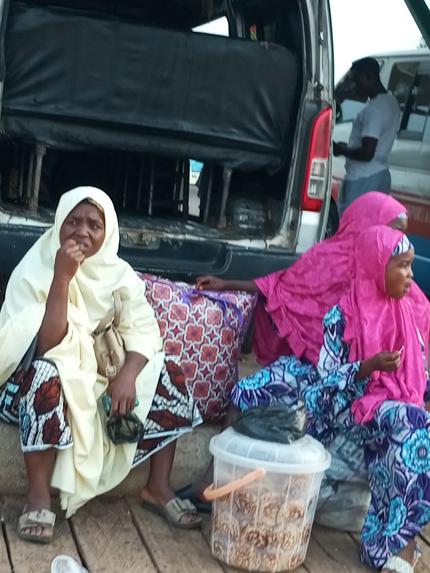 In Gombe, Nigeria, three individuals are gathered near the open rear of a vehicle. On the left, a woman wearing a cream-colored headscarf and patterned skirt sits with her hand to her mouth, appearing to be eating. She has a beige handbag slung over her shoulder. To her right, a younger person in a bright pink hijab and a patterned blue and purple outfit is seated. Another person in a similar pink hijab is partially visible behind them. In front of the seated individuals is a transparent plastic bucket filled with what appear to be pastries or biscuits, covered with a black plastic bag. Behind them, a man in a white t-shirt and light-colored pants stands and looks down at his phone. The scene is set on a wooden surface, possibly a dock or a makeshift platform, with a large vehicle, its back open, dominating the background.