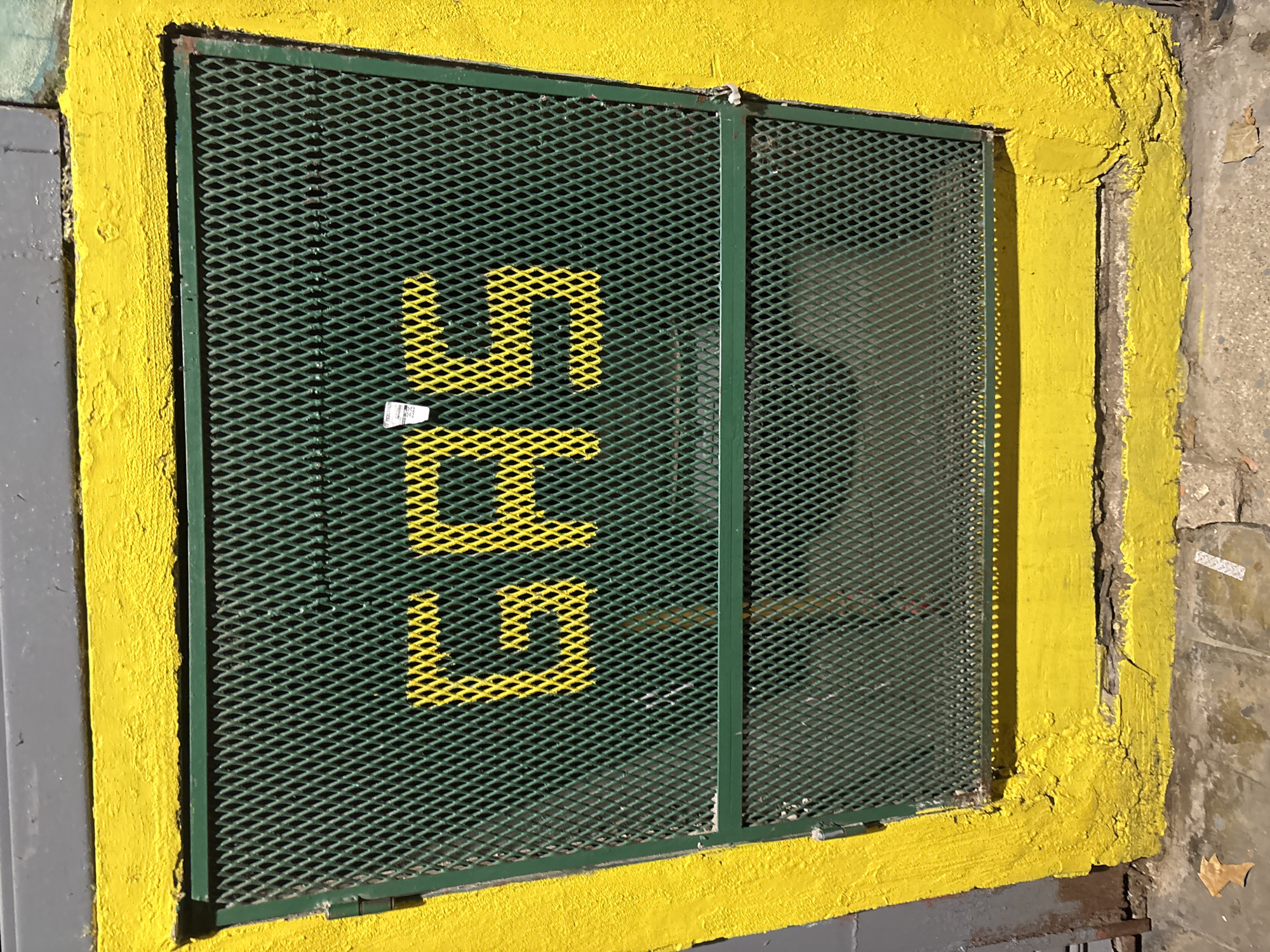 The image displays a prominent, bright yellow-painted rectangular frame surrounding a dark green metal mesh grate, likely covering a window or ventilation opening on an outdoor wall. The scene is set in an urban environment, possibly Buenos Aires, Argentina, under bright daylight conditions, indicated by strong shadows cast by the mesh onto an interior surface.
The green grate is made of expanded metal mesh, exhibiting a diamond pattern, and is divided vertically by a slim, dark green metal bar. Bold, blocky letters "LLL" are painted in yellow directly onto the left section of the green mesh, arranged vertically. A small, white rectangular sticker or label is affixed to the mesh near the center of the "LLL" text, but its details are illegible.
The vibrant yellow frame surrounding the grate appears to be made of concrete or stucco and shows signs of wear, with chipped paint and a rough, textured surface, particularly visible along its edges and on the right side where underlying concrete is exposed. The wall adjacent to the yellow frame on both the left and right is a dull, unpainted grey, providing a stark contrast to the bright yellow. No people or specific activities are depicted; the image captures a static architectural detail. The overall impression is one of a functional structure given a striking, if somewhat aged, aesthetic treatment.