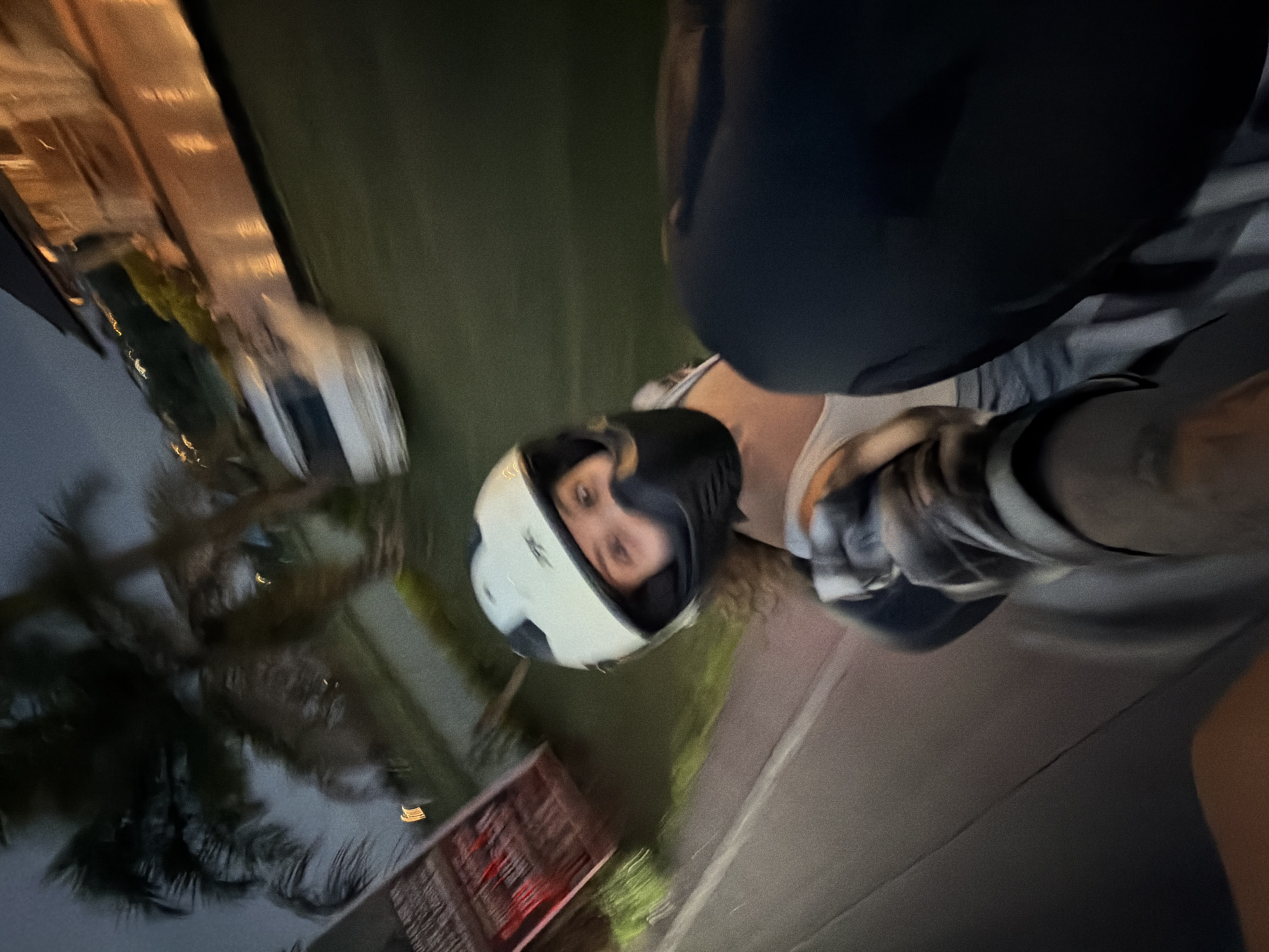A person wearing a white helmet with a black visor is riding on a scooter, seen from a tilted perspective. The individual has brown hair and fair skin. The person behind them is wearing a dark helmet and a gray plaid shirt. The scooter is moving on a road, and in the background, there are trees, a white car, and buildings. The photo was taken in North Kuta, Indonesia.