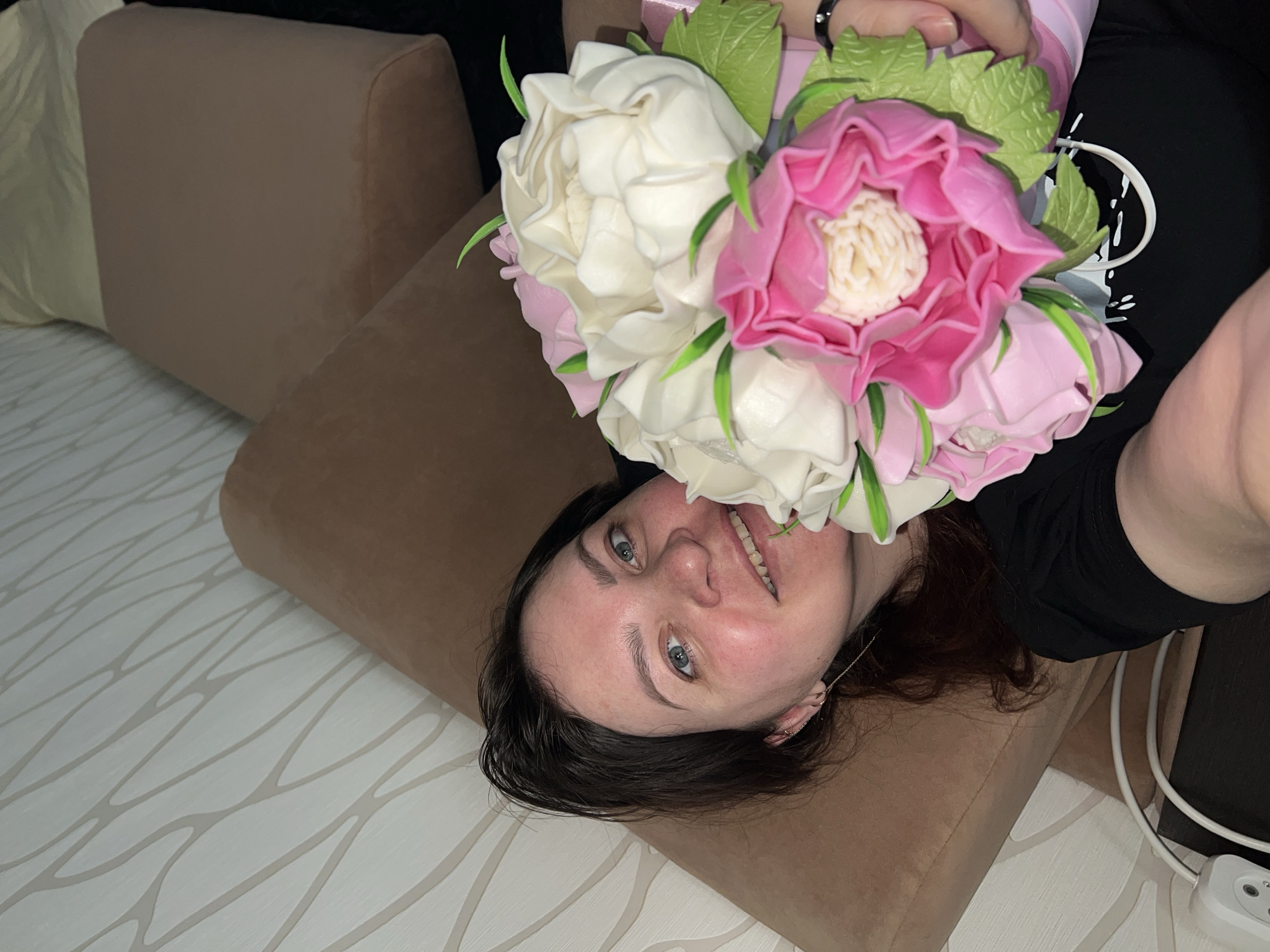 A woman is lying on a taupe pillow on a white patterned surface. She holds a bouquet of white and pink flowers in front of her face. She has blue eyes and dark hair. The background includes a brown couch and black furniture. The photo was likely taken indoors. The location cannot be determined from the image.