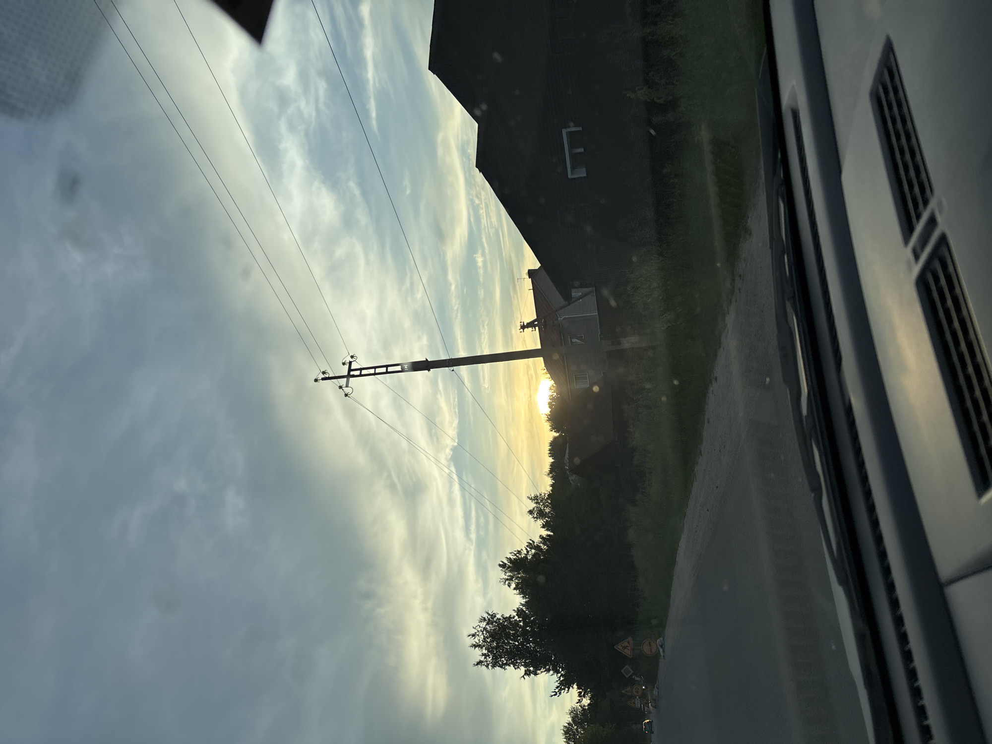 The image depicts a rural roadside scene with a dramatic sky at sunset, viewed from inside a vehicle. The perspective suggests the camera is positioned on the dashboard, with the windshield in the foreground. The sky is a blend of blues, grays, and yellows, with wispy clouds streaked across it. The setting sun is a bright, hazy orb peeking through the clouds, casting a warm glow on the scene.
On the right side of the image, a dark, angular house with a prominent pitched roof dominates the middle ground. A single, rectangular white window is visible on its dark facade. To the left of this house, a utility pole with multiple wires stretches vertically into the sky, bisecting the frame. Further left, a dense line of trees forms a dark silhouette against the brightening sky.
In the foreground on the left, a paved road curves gently to the left, disappearing into the distance. Several traffic signs are visible along the side of the road, including caution signs with yellow triangular shapes. A single vehicle is partially visible on the road in the far distance. The grassy verge beside the road is overgrown with weeds and wildflowers. The overall impression is of a quiet, possibly remote, location bathed in the soft, diffused light of dusk. The scene is devoid of any identifiable human figures. The location is identified as Chigiri, Russia.