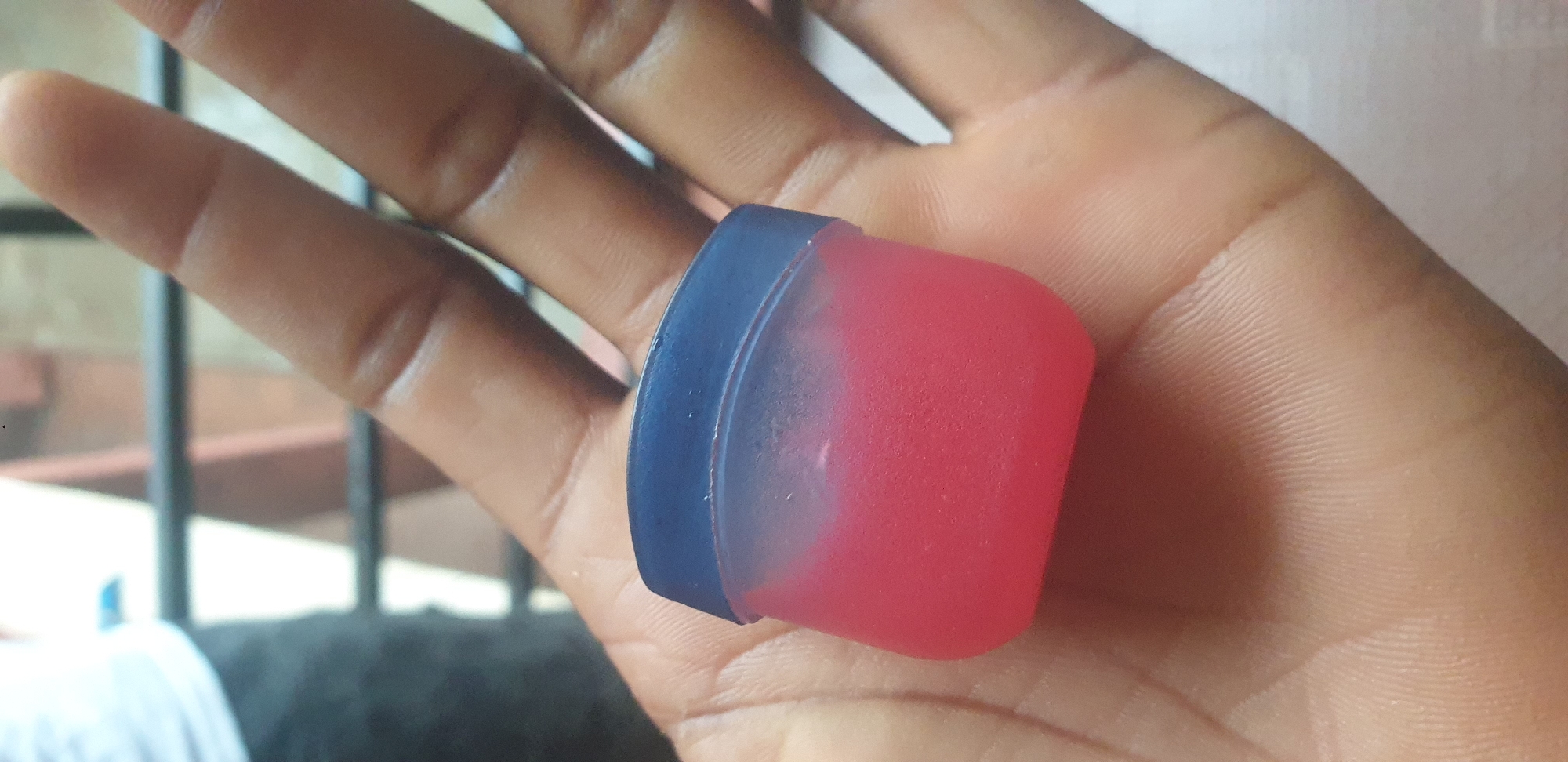 In Yelwa, Nigeria, a person's open hand, likely held in a slight cupping gesture, supports a small, square, translucent plastic container with a dark blue lid. The container is filled with a vibrant pink substance, possibly a lip balm or a similar cosmetic product, which appears somewhat granular. The skin of the hand is dark-skinned, with visible fingerprints and creases on the palm and fingers. In the blurred background, a metallic railing and indistinct figures can be observed, suggesting an outdoor or semi-enclosed urban setting.
