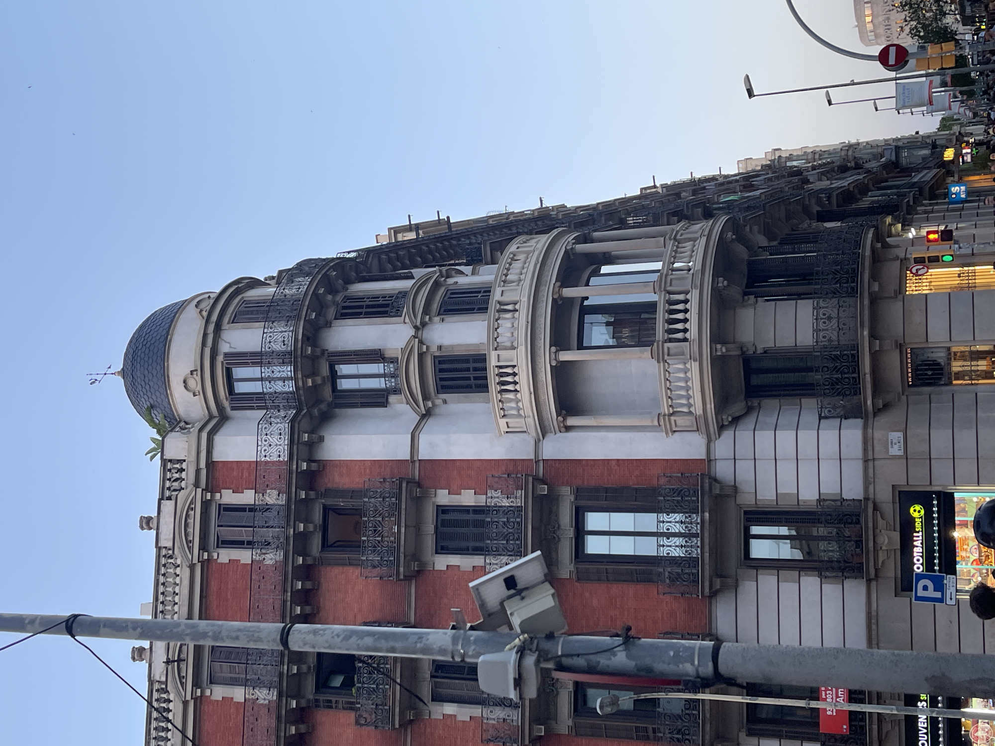This is a medium-angle, full-shot, outdoor photograph taken in Barcelona, Spain. The composition focuses on a corner building with distinct architectural features. The building on the left has a red brick facade with cream-colored trim around the windows and balconies. It features dark wooden shutters on the windows and ornate wrought-iron balconies with decorative grills. To the right, the building transitions to a cream-colored facade, also with similar windows and balconies, culminating in a rounded corner topped by a dark dome with a weather vane. There are multiple stories visible, with balconies on each level. A gray utility pole with electrical equipment is prominently positioned in the foreground on the left side of the frame. The sky is clear and blue with a few faint birds visible. On the street level, there are shopfronts with signage, including one that says "FOOTBALL SIDE" with a soccer ball logo. Traffic lights and street signs are also present. The overall impression is an urban streetscape with a focus on a historical architectural style.