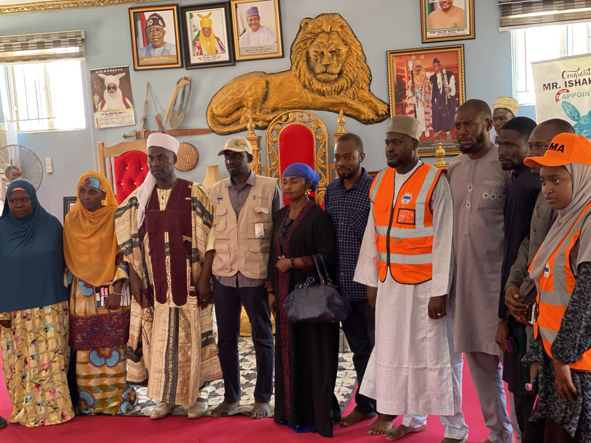 A group of about a dozen people are gathered on a red carpet in an indoor setting, possibly a government building or royal residence, as indicated by the presence of several framed portraits of public figures and a large golden lion sculpture above an ornate red throne. Some individuals wear traditional African clothing, while others don modern attire, including a few wearing bright orange safety vests and caps. A woman in the group carries a dark-colored handbag. The location is suggested by the backdrop and attire, possibly in Sabon Tasha, Nigeria. A banner congratulates "Mr. Ishak" on their appointment. The individuals appear to be posing for a photograph.