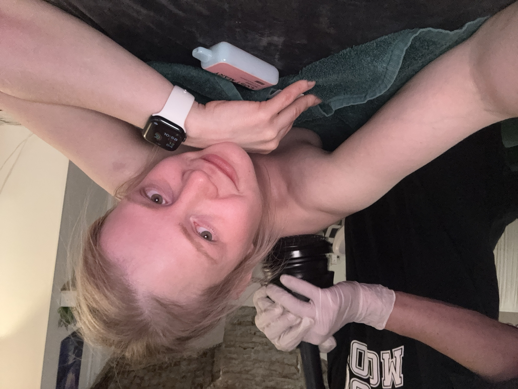 A woman is reclining on a dark, possibly upholstered surface, her arms positioned above her head. She is wearing a white smartwatch on her left wrist. A blue and pink bottle is resting on a folded green towel near her. Another person, wearing white latex gloves and a black t-shirt with white lettering, is holding a black cylindrical object. The location is likely indoors, with a glimpse of a textured wall and some furniture visible in the background. The location in the city of Saint Petersburg, Russia, cannot be determined from the provided image.