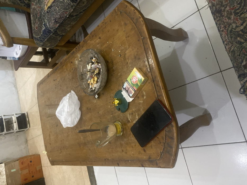 The image depicts an indoor scene within a living space in Cimenyan, Indonesia. A rectangular wooden coffee table, exhibiting surface wear and scratches, occupies the central foreground. On its surface, from left to right, are: a clear glass mug containing a small amount of light brown liquid and a silver spoon; a crumpled white plastic bag; and a clear glass ashtray filled with numerous cigarette butts, ash, and discarded foil. To the lower left of the ashtray, a red-backed smartphone with a dark, inactive screen lies face-up. Adjacent to the smartphone is a green cigarette pack, identifiable as "Gudang Garam 76," and behind it, a small green coaster topped with a golden crown-shaped decorative item. A small silver-colored rectangular object rests near the ashtray. The floor consists of large white tiles with dark grout lines. In the background, to the left, a brown cardboard box is visible against a light-colored wall. Towards the center-back, a white, multi-tiered shoe rack holds several pairs of casual footwear. To the right, part of a wooden armchair or sofa with patterned upholstery is visible, and behind it, a small wooden side table supports a white appliance, possibly a water dispenser. No individuals are present in the scene.