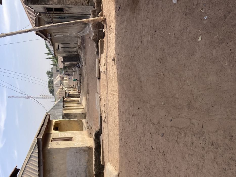The image depicts a residential street in Minna, Nigeria, under clear daytime conditions. The foreground features an unpaved, reddish-brown dirt road with visible tire tracks and scattered debris. Two horizontal concrete slabs or blocks are positioned across the road in the immediate foreground. Single-story buildings with light-colored, weathered walls and corrugated metal roofs line both sides of the street, receding into the distance.
On the left, a house with a brown window frame and an arched entryway is partially visible. On the right, a wooden utility pole stands prominently, supporting overhead wires that extend down the street. In the mid-ground, a person wearing a green shirt walks away from the viewer, while other indistinct figures, including what appear to be children and small dark animals (possibly chickens), are visible further down the road.
The street slopes gently downward towards the background, where more residential structures are present. A tall, red and white striped lattice communications tower stands against the light blue sky in the distant background, with additional overhead wires visible beneath it. Shadows are distinct, consistent with direct sunlight.