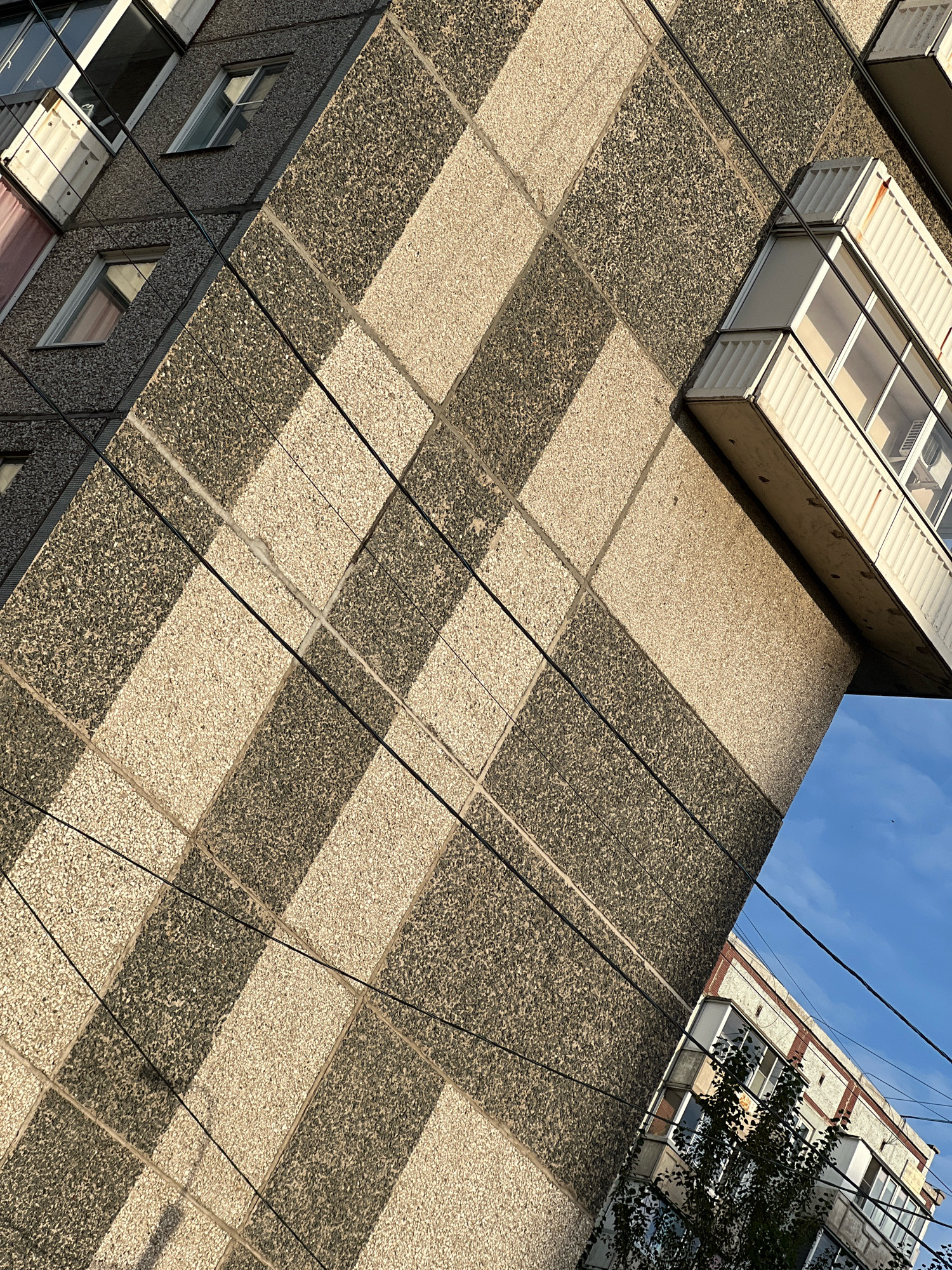The image shows a close-up of a grey and beige tiled building facade in the city of Krasnoyarsk, Russia. The tiles are arranged in a diagonal, checkered pattern, with shadows cast by the sun creating a further geometric effect. Several windows and a balcony are visible on the building. Thin black electrical wires crisscross the frame. In the lower right corner, a portion of another building with red and white trim is visible, along with some green foliage. The sky above is blue with some clouds. The overall impression is of a residential building with a distinctive architectural style, likely from the Soviet era.