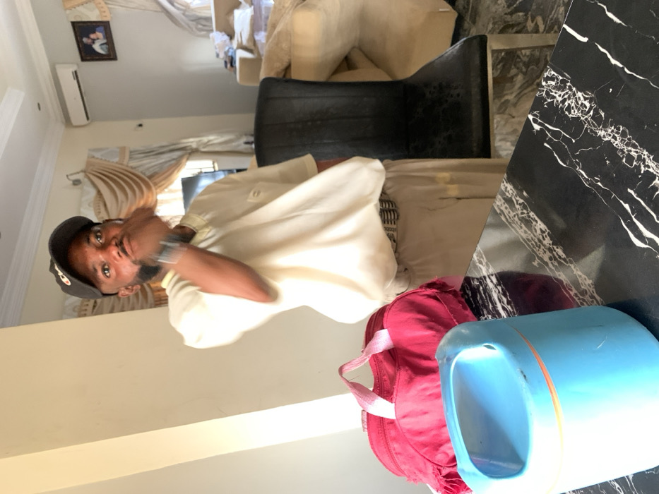 A person wearing a hat and a light-colored shirt is indoors. A black chair is near the person. A red bag and a blue container are in the lower right corner. In the background, there's a curtained window and a cream-colored sofa. A small framed picture is on the wall. The image appears to be rotated.