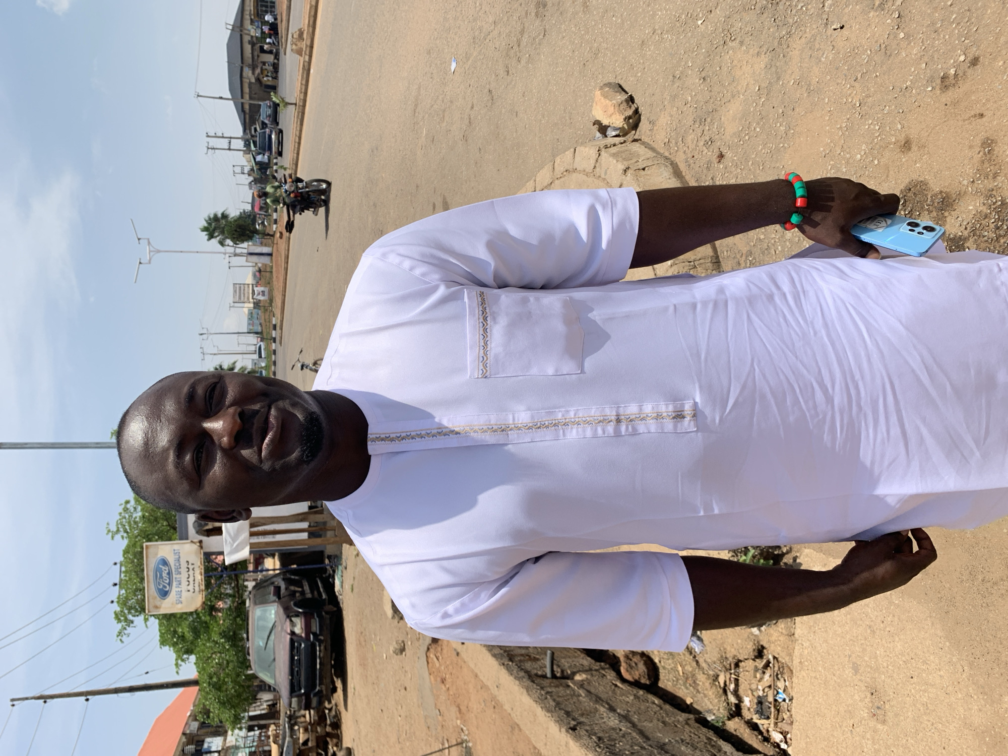 A man stands on a street in Rafin Par, Nigeria, holding a blue phone. He is wearing a white traditional shirt with decorative trim around the pocket and neckline. He also wears a bracelet with red and green beads. Behind him are vehicles, buildings, and power lines. A Ford spare parts shop sign is visible in the background. The street is paved and appears to be moderately busy. A motorcyclist can be seen riding in the distance.