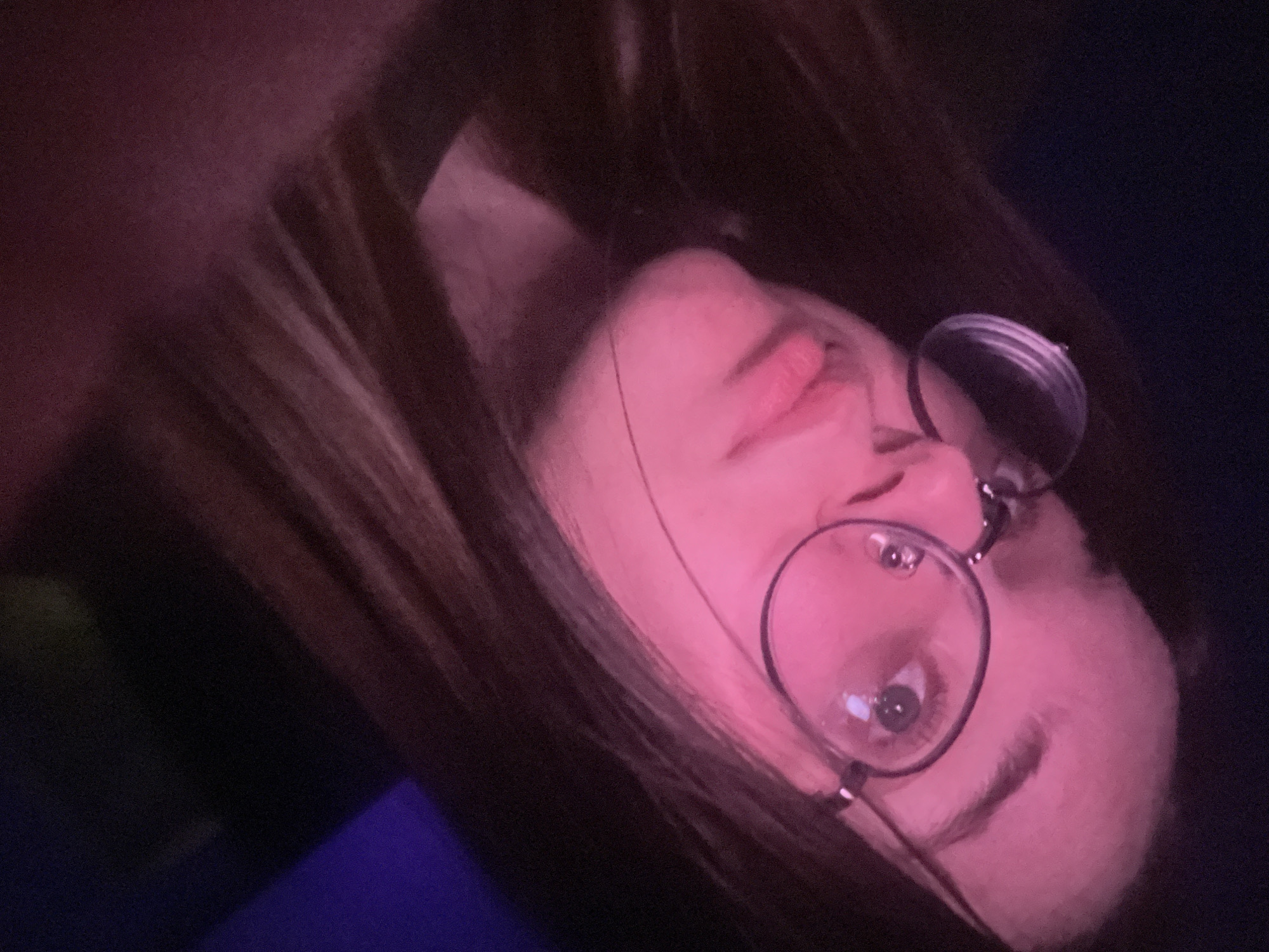 A close-up, low-angle portrait features an individual, likely female, with long dark hair and pale skin, illuminated primarily by a strong pink/magenta light source from the left. The subject is wearing round, clear-rimmed glasses with dark metal frames, through which their right eye (viewer's left) is visible looking upwards and slightly to the right. The mouth is closed with a slight pout. The pink light heavily colors the subject's face, neck, and portions of the hair. The background is dark and indistinct, with a muted purple area visible in the upper right quadrant. The specific location within the city of Pushchino, Russia, cannot be determined from the visual information presented in this image.