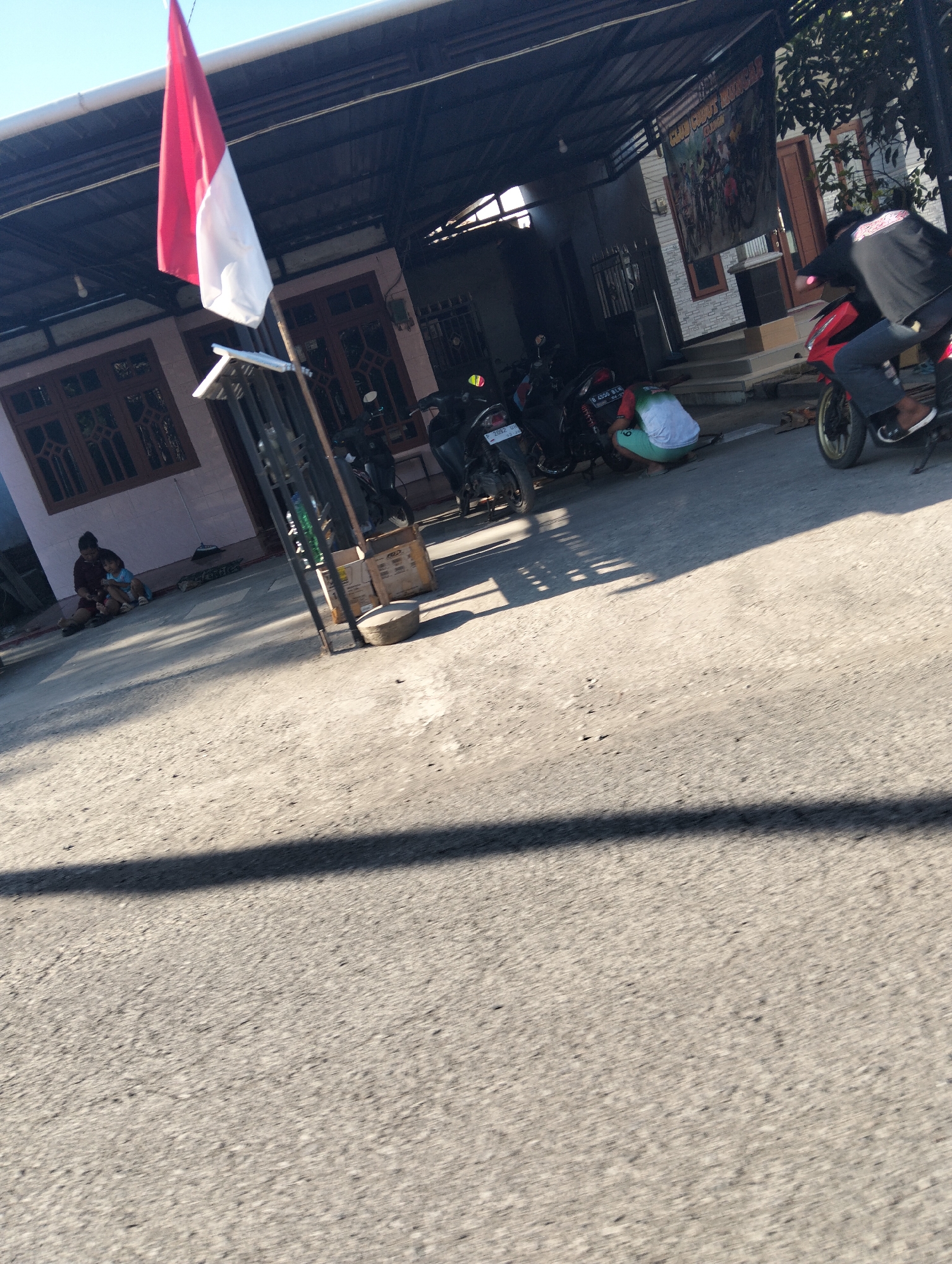 The image depicts a scene in front of a building with a dark-colored roof and a light-colored facade. A red and white flag, likely the Indonesian flag, is visible, hanging from a pole. Several motorcycles are parked in front of the building. In the foreground, there is a road with a distinct shadow cast across it.

In the left portion of the image, two individuals, an adult and a child, are seated on the ground. Towards the center and right, there are more individuals. One person is crouched down, seemingly working on or near a motorcycle. Another person is riding a red motorcycle, with their back to the camera. A banner with text and images of people on bicycles hangs on the building in the background. The overall environment appears to be an outdoor commercial or residential area.

The location is identified as Kedungringin, Indonesia, suggesting a street-level view of a community or business area. The lighting indicates it is daytime, with sunlight casting shadows.