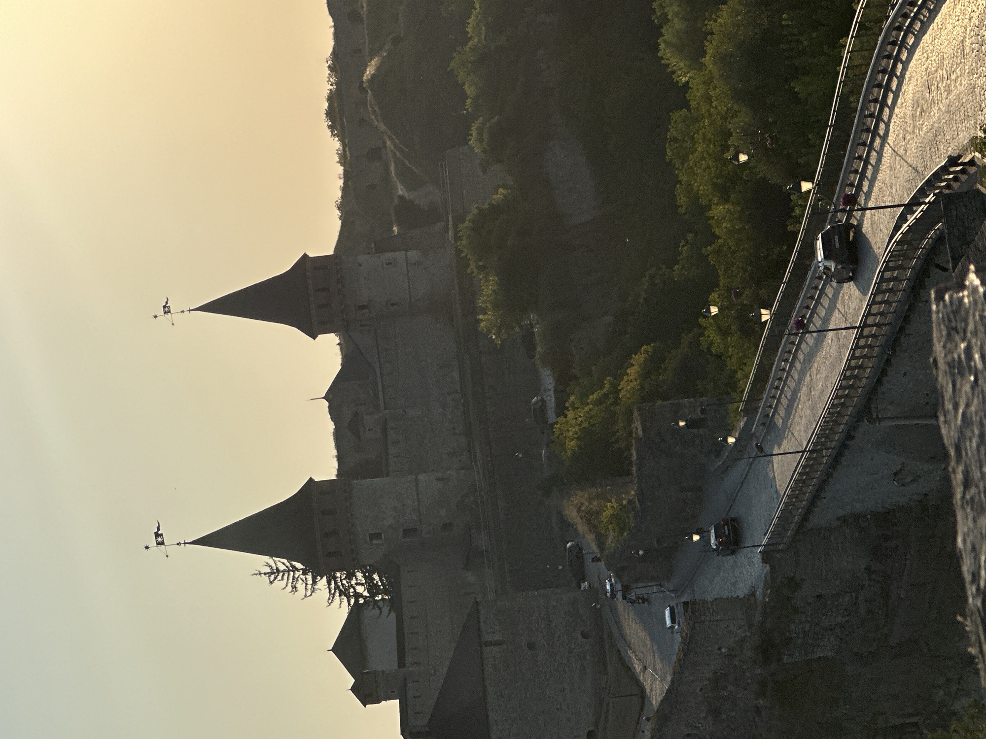 The image depicts the Kamianets-Podilskyi Castle in Kamianets-Podilskyi, Ukraine, at dusk. The castle, constructed of stone, features two prominent conical towers, each topped with a flag. A winding cobblestone road leads up to the castle, and a bridge with decorative lamps crosses a gorge in the foreground. Several cars are visible on the road and bridge. Lush green trees and vegetation surround the castle and the gorge. The sky is a pale orange and yellow, indicating the setting sun. The overall scene is a picturesque view of the historic fortress and its surrounding landscape.