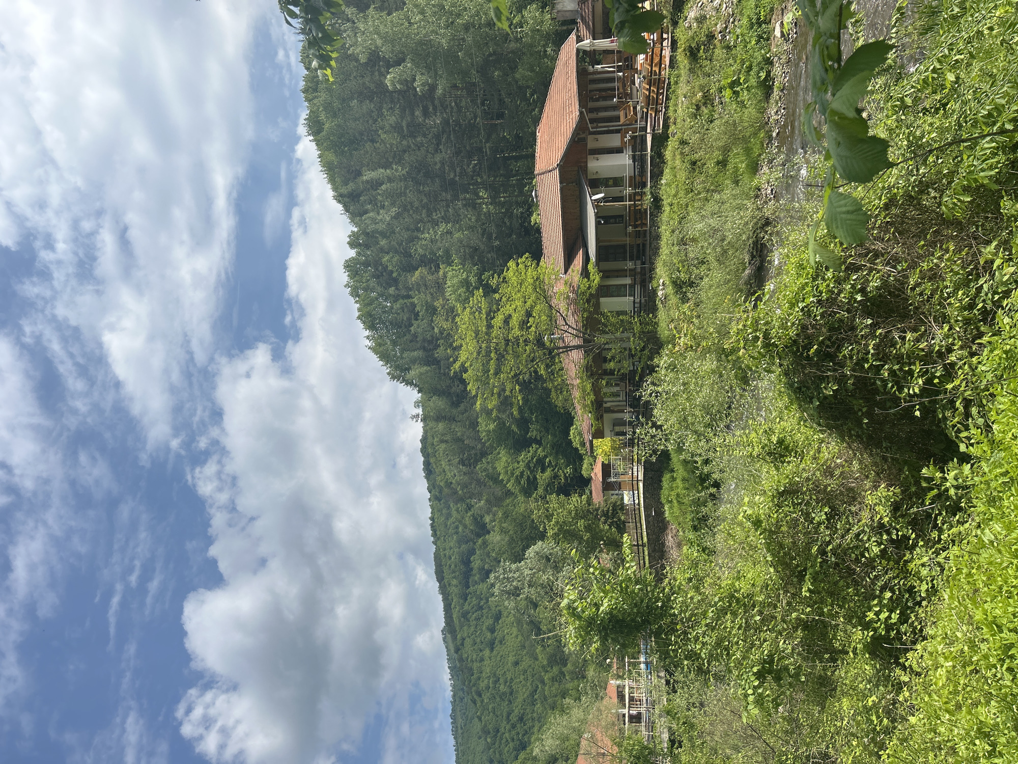 The image depicts a lush, green landscape with a building nestled into a hillside. The building, which appears to be a multi-story structure with a red-tiled roof, is situated alongside a stream or river that runs parallel to it. Dense foliage and trees cover the hillside, with the sky visible above featuring a mix of blue and white clouds. The precise location within the city of Unknown, Unknown, cannot be determined from the provided image.