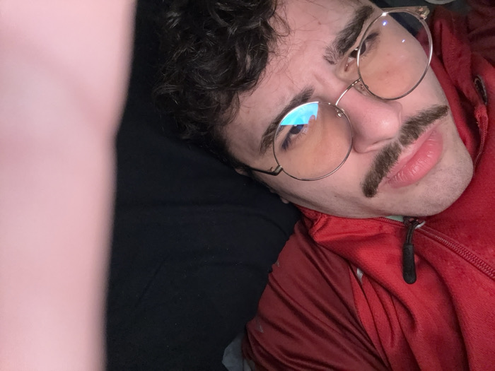 A person with dark curly hair and a prominent mustache is reclining. They are wearing round gold-rimmed glasses and a red zippered jacket. The person appears to be lying on a black surface, possibly a couch or bed. A blurry pink shape is visible on the left side of the frame, possibly an arm or leg close to the camera. The individual's expression is slightly quizzical or skeptical.