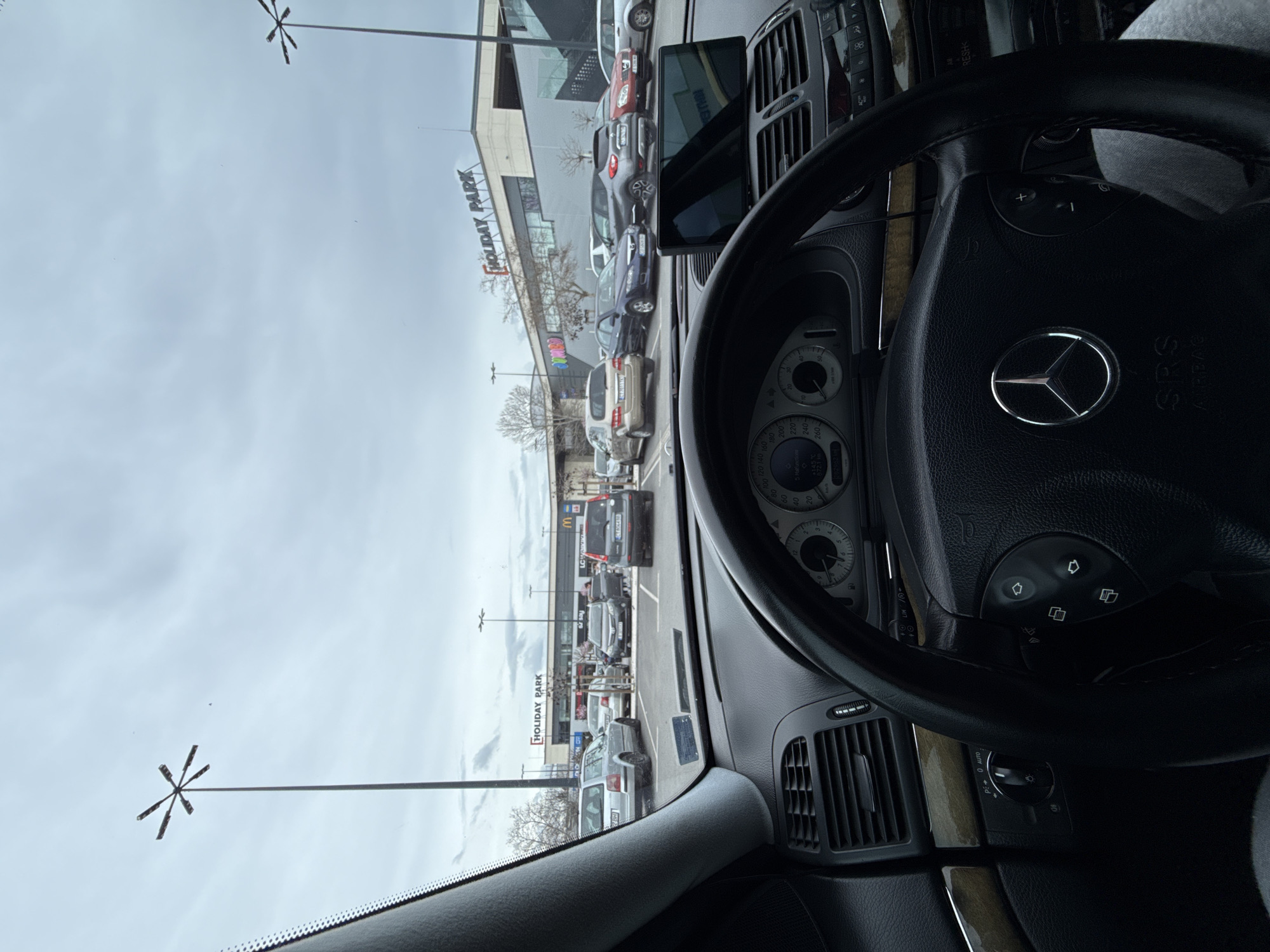 The image depicts a first-person view from inside a dark-colored Mercedes-Benz vehicle, specifically an E-Class (W211 generation), looking out into a parking lot on an overcast day. The steering wheel, dashboard, and instrument cluster are visible in the foreground, with a silver Mercedes-Benz logo centered on the steering wheel and an "SRS AIRBAG" label below it. A separate electronic screen device is mounted on the central air vents above the car's built-in infotainment system.
Beyond the vehicle's windshield, a large outdoor parking area is visible, occupied by numerous parked cars of various makes and models, including a silver Volkswagen Passat station wagon, a dark grey Volvo, and several other sedans and SUVs. Parking lines delineate the spaces. In the background, a large commercial building is present, identified by a prominent sign that reads "HOLIDAY PARK." Below "HOLIDAY PARK," a McDonald's logo and "LC Waikiki" are visible, with "JUMBO" partially visible on the left side of the building. Tall, distinctive streetlights with multi-directional luminaires are scattered throughout the parking lot and in the distance. The sky is uniformly gray and overcast, and leafless trees are visible in front of the commercial building, suggesting a cold season. This scene is located at Holiday Park in Stara Zagora, Bulgaria.