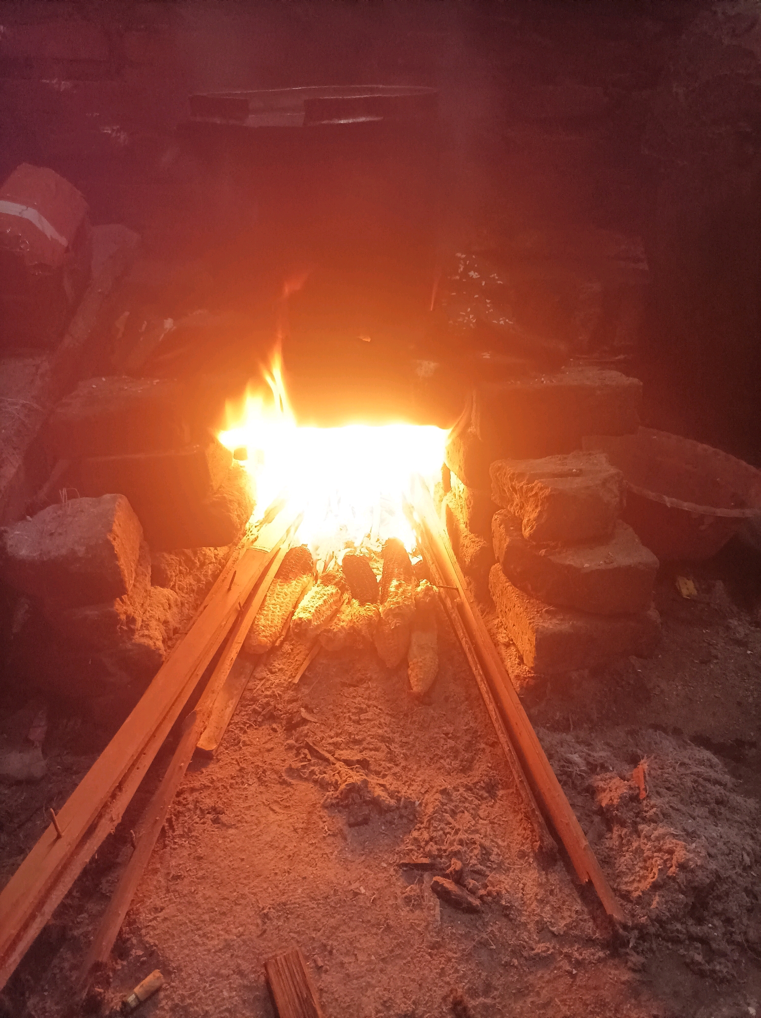The image shows a close-up view of a traditional oven in Canga'an, Indonesia, in operation. A bright fire burns in the center, illuminating the surrounding area with a warm orange glow. The oven appears to be constructed from bricks, with some stacked on the sides. Several pieces of wood are placed near the fire, possibly for fuel or to support items being cooked. On the ground, there are also what appear to be ears of corn, suggesting food is being prepared. The ground is covered in a mixture of dirt and ash, indicative of a cooking environment. The overall impression is of a rustic and functional setup for outdoor cooking.