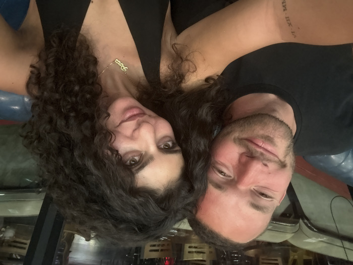 A close-up photograph shows two individuals, one male and one female, seated together indoors within what appears to be a restaurant or bar setting in Los Angeles, United States of America.
The male, positioned on the left, has light skin, short brown hair, and stubble. He wears a black crew-neck t-shirt. A tattoo displaying Roman numerals "VI-XII-MXX" is visible on his left forearm. His forehead and nose exhibit some redness.
The female, positioned on the right, has olive skin and long, dark, curly hair. She wears a black v-neck top and a gold-colored necklace with a "Paris" pendant.
Both individuals are smiling and looking directly at the camera. They are seated on a blue tufted bench or booth, partially visible in the foreground. The background is blurred, revealing dim lighting and indistinct wooden structures, consistent with an interior establishment. The image provides no specific visual cues to identify the exact location within Los Angeles.