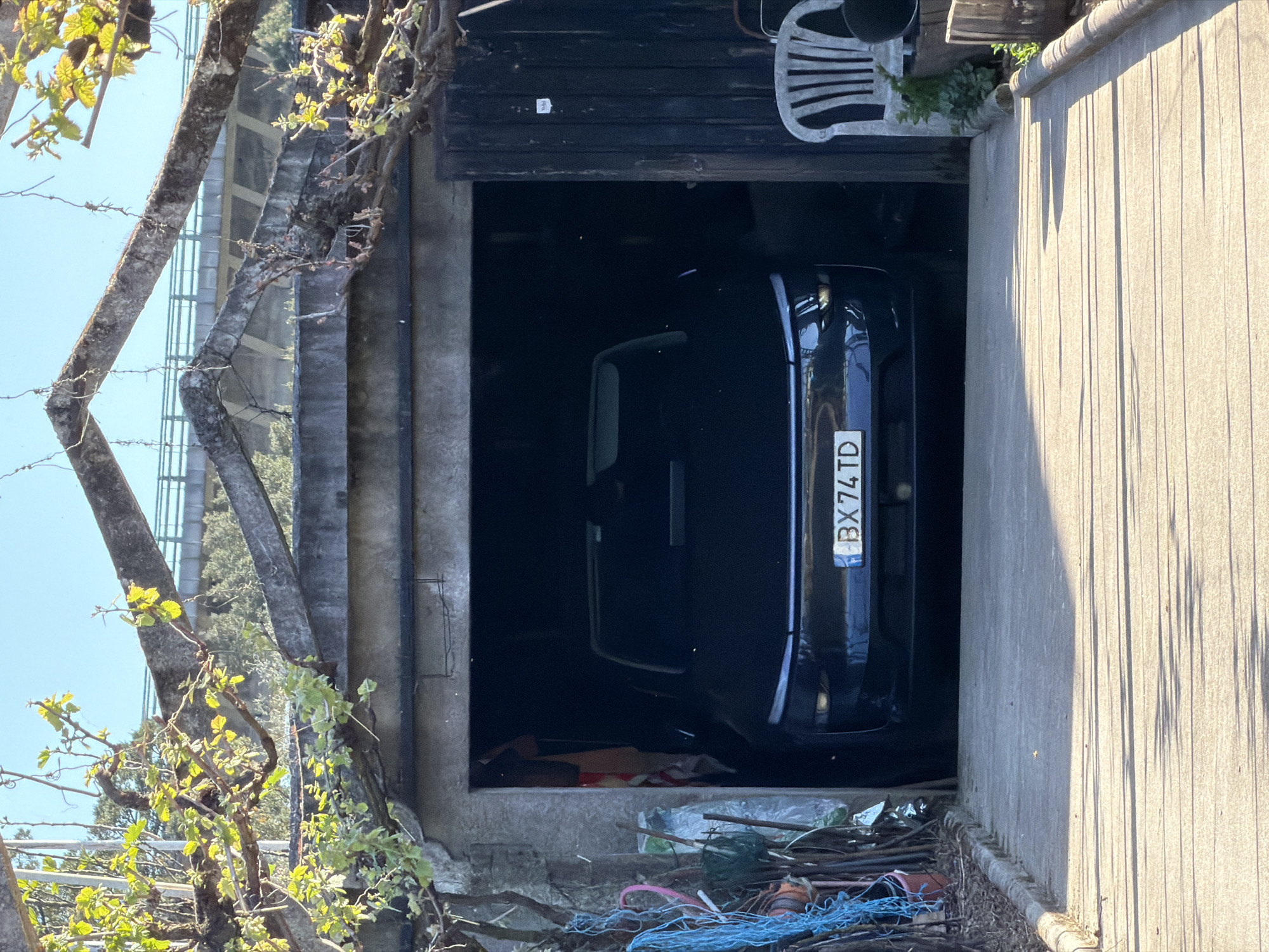 The image shows a dark blue car parked inside a garage. The car's license plate, "BX 74 TD," is visible. The garage is located between a concrete wall and a stone wall. The entrance to the garage is partially obscured by branches and foliage. In the area around the garage entrance, there are various items including a white plastic chair, debris, and some ropes. The setting appears to be in Vila Real, Portugal, based on the license plate format, which indicates a Portuguese vehicle registration.