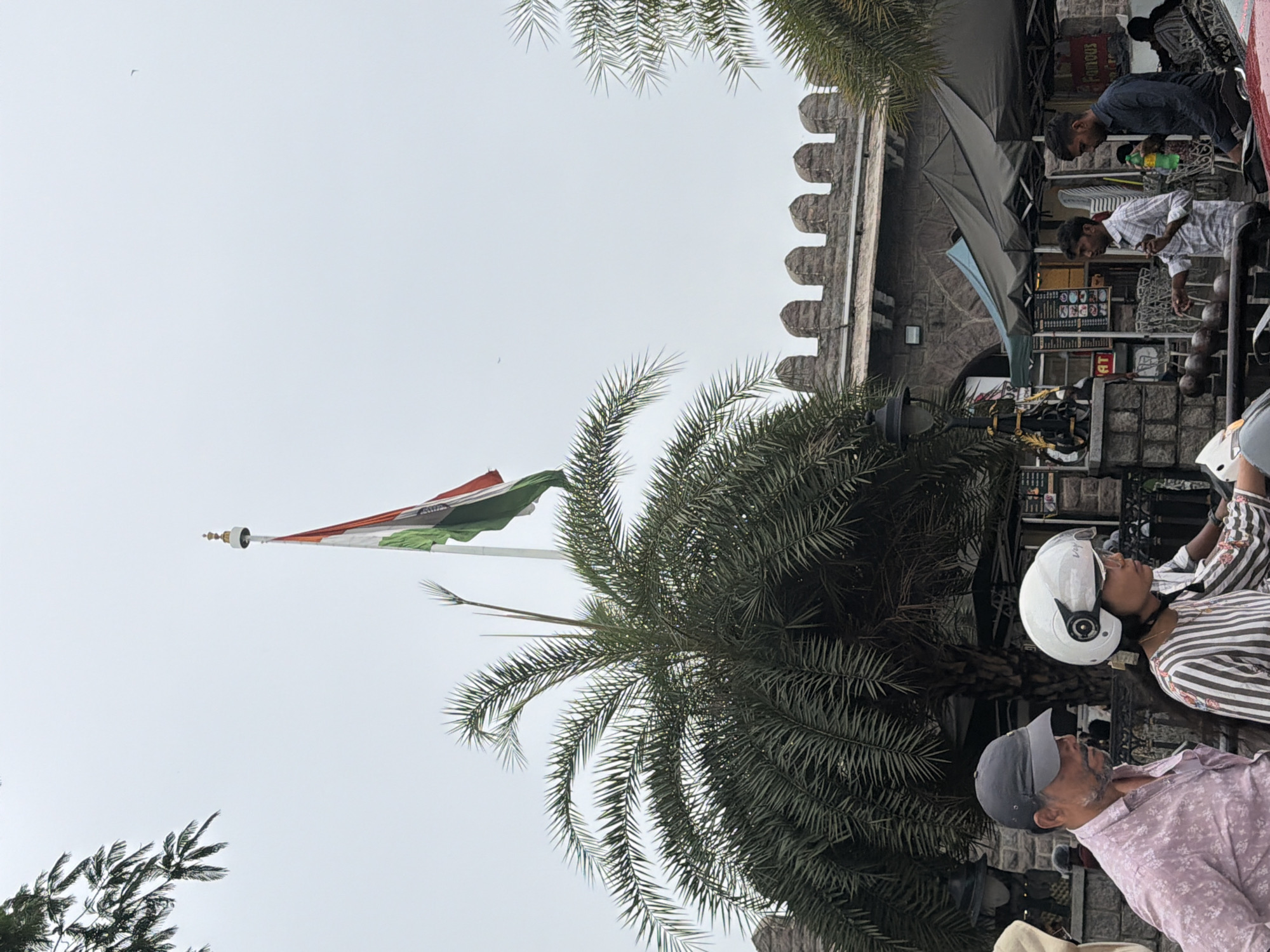 In this image, an Indian flag is hoisted on a flagpole against a cloudy sky. In the foreground, a woman wearing a white helmet rides a scooter, and a man in a pink patterned shirt and a grey baseball cap stands beside her. Palm trees are visible on the left and right sides of the image. Behind them, a stone structure with crenellated battlements can be seen, possibly part of a fort or historical building. Several tents are set up in front of the structure, with people visible in and around them. The scene appears to be a public space in Hyderabad, India, likely near a landmark.