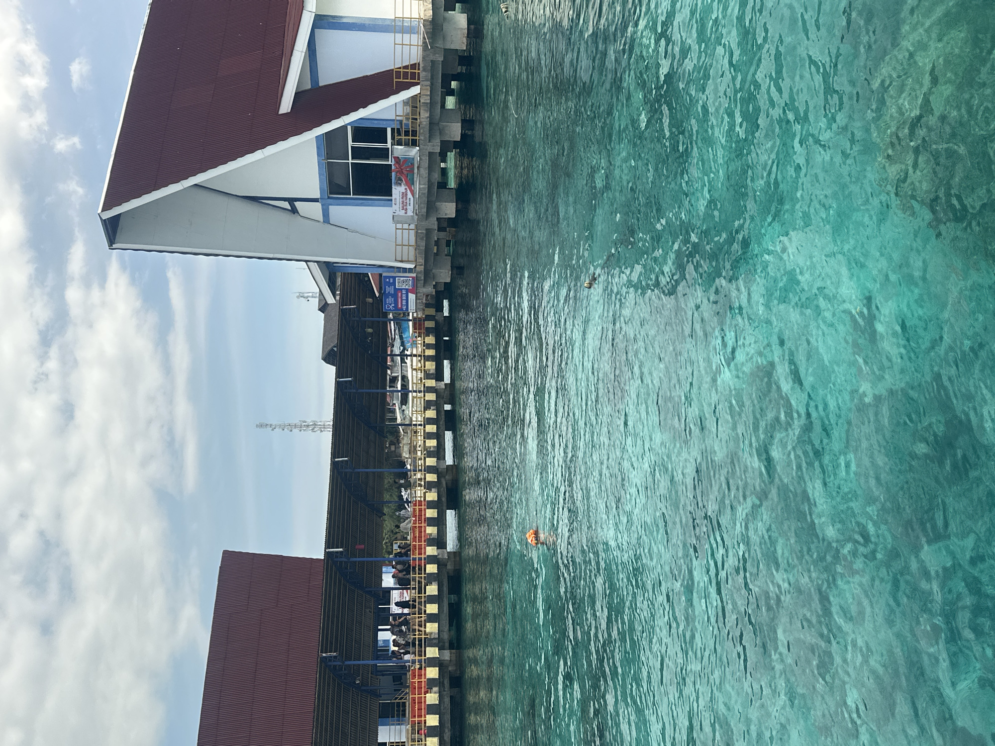 The image shows a waterfront scene. There is a building with a red roof and white walls with blue accents along a pier. The pier is made of dark colored concrete. There are people seated underneath the pier and along the railing. The water appears turquoise in color, and there is a person wearing an orange floatation device in the water. The location is Unknown, Indonesia.
