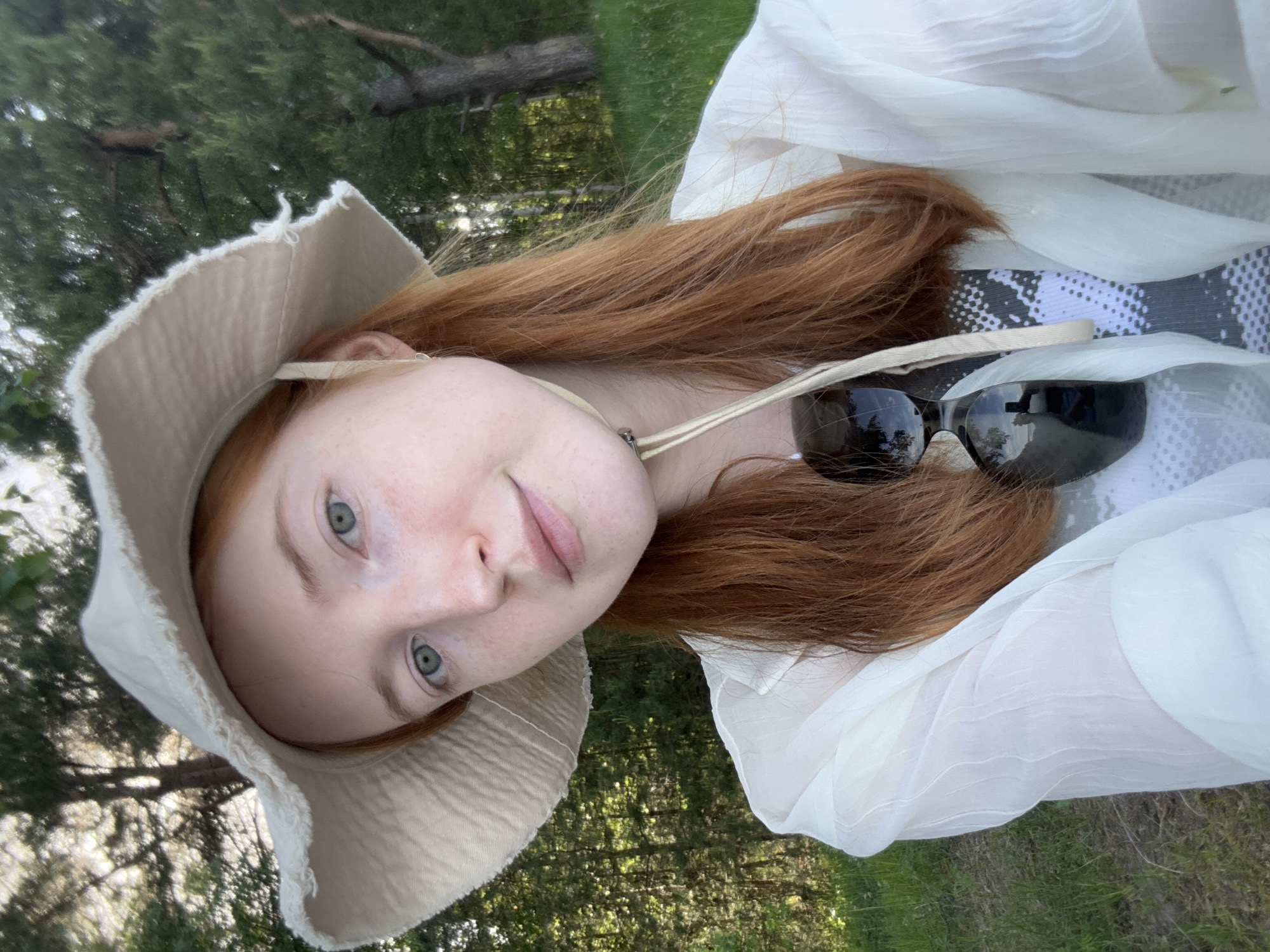 A young, fair-skinned woman with red hair and light-colored eyes poses for a selfie outdoors. She wears a wide-brimmed, light-colored hat with a frayed edge. Sunglasses rest on her red hair, and she wears a lightweight white blouse over a black and white patterned top. Green grass and trees are visible in the background, partially obscured by the camera angle. It is not possible to confirm the location is Chaikovsky, Russia, based on the visual information provided.