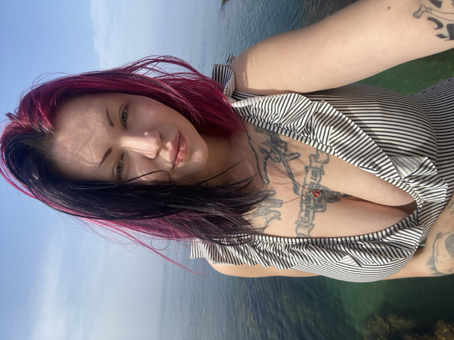 The image depicts an adult female in a close-up selfie format, positioned outdoors in Kunje, Montenegro, with a body of water in the background. She has light skin and bi-colored hair, centrally parted, with the left side dark (black/dark brown) and the right side magenta. Her hair appears tousled. She looks directly at the camera with a neutral facial expression.
She wears a black and white striped sleeveless top or swimsuit featuring ruffled straps and a deep V-neckline. Multiple tattoos are visible on her upper chest and arms. A prominent chest tattoo is a banner containing text that partially reads "Everything you can". A silver chain necklace with a red, heart-shaped pendant rests on her chest.
The background consists of a clear blue sky meeting a calm expanse of sea or ocean. The water directly behind her exhibits a clear, greenish hue, revealing submerged dark rocks beneath the surface. The scene is illuminated by bright daylight.