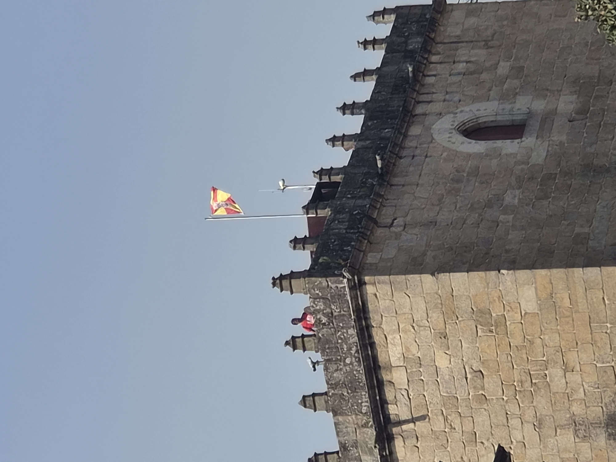 The image shows a view of the ramparts of a stone tower in Barcelos, Portugal. A person wearing a red shirt is visible on the ramparts. A flag, which appears to be the flag of Galicia, is flying from a flagpole on top of the tower. The sky is clear and blue. The tower is made of rough-hewn stone blocks and has decorative crenellations along the top. A gothic-style arched window is visible on the side of the tower.