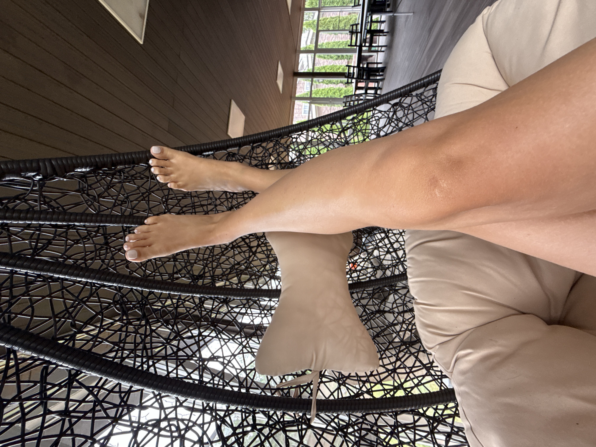 A person's legs are crossed and raised in a hanging chair, with their feet visible at the top of the frame. The chair is made of black woven material and is suspended from the ceiling. A light-colored pillow is placed behind the person's legs. The background shows a room with wooden paneling on the ceiling and a row of windows looking out onto a green, outdoor space with trees and tables and chairs. The location is not specified as being in the city of Kommunarka, Russia.