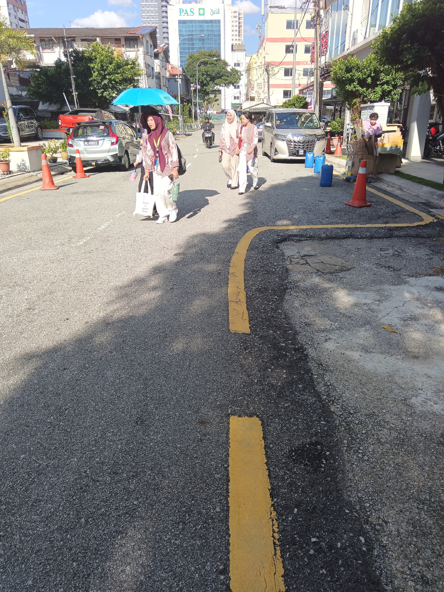 The image depicts a street scene in Kuala Lumpur, Malaysia, likely in a commercial or residential area with buildings lining the road. In the foreground, a paved road with yellow markings is visible, with some patches of wear and tear. Several people are present. On the left, a woman in a hijab walks under a bright blue umbrella, carrying shopping bags. To her right, two other women, also in hijabs, are walking. In the middle of the road, a silver minivan is parked. Further down the street, a man is pushing a cart laden with boxes and blue barrels, with an orange traffic cone nearby. A motorcycle is also visible further down the street, with a rider. The background shows a mix of buildings, including some older structures and a modern, multi-story building with the "PAS" logo. Trees and some potted plants are present along the roadside. The sky is blue with some clouds, indicating daylight.