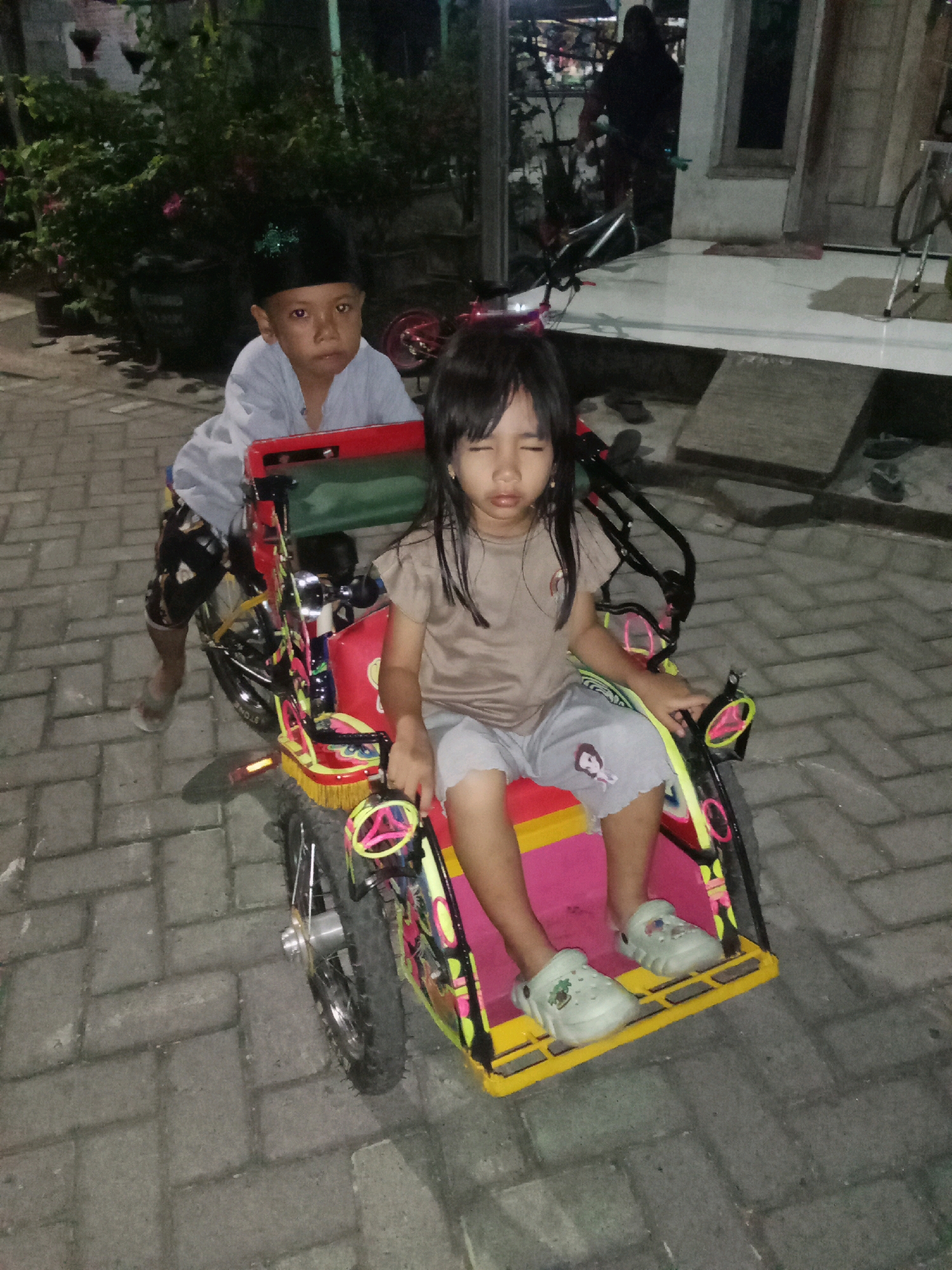 In Sidorejo, Indonesia, two children are pictured at night, seated on a colorful, toy-like tricycle designed for passengers. The tricycle is red and yellow, with bright green and pink accents and decorative tassels. A young boy, wearing a black cap and a light blue long-sleeved shirt, is positioned behind the tricycle, with his legs dangling on either side of the frame. He appears to be pushing or pulling the vehicle, with one foot on the ground. In the passenger seat is a young girl wearing a light brown t-shirt and grey shorts adorned with cartoon characters. She is looking down with her eyes closed. Both children are wearing light-colored, slip-on shoes. The tricycle sits on a paved brick walkway. In the background, lush green foliage and potted plants are visible, suggesting a residential area. A building with a white tiled porch and a dark doorway is also present. Another person, whose face is not visible, is walking in the background, carrying a bicycle. The scene is illuminated by artificial light, creating shadows on the ground.