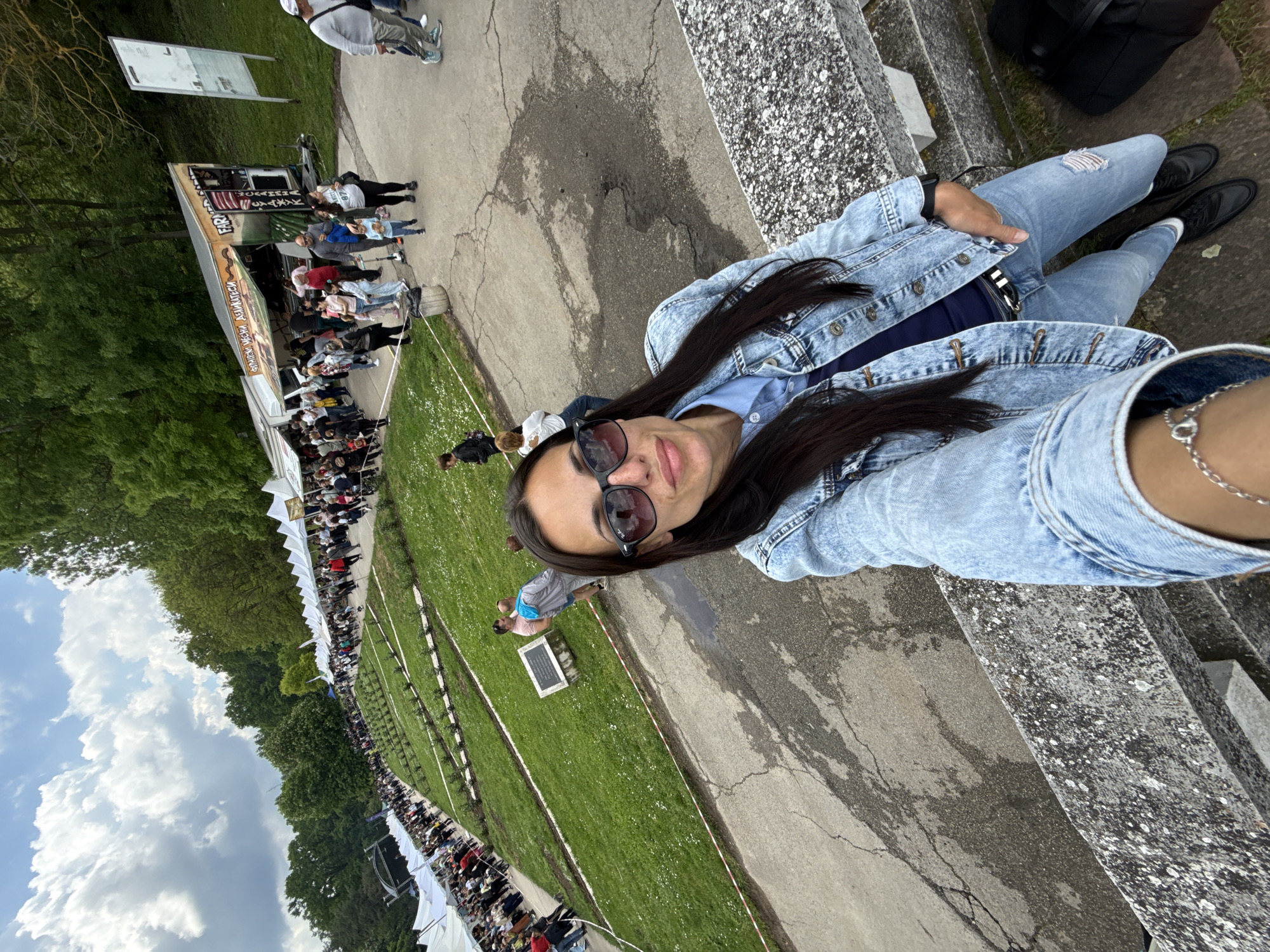 The image is a selfie of a woman wearing sunglasses and a denim jacket, taken outdoors in Pleven, Bulgaria. She is sitting on a concrete structure. In the background, there's a large gathering of people, likely attending an event with white tents set up. A food stall is visible. The ground is a mix of concrete walkways and grassy areas. Trees are visible in the background. The sky is overcast.