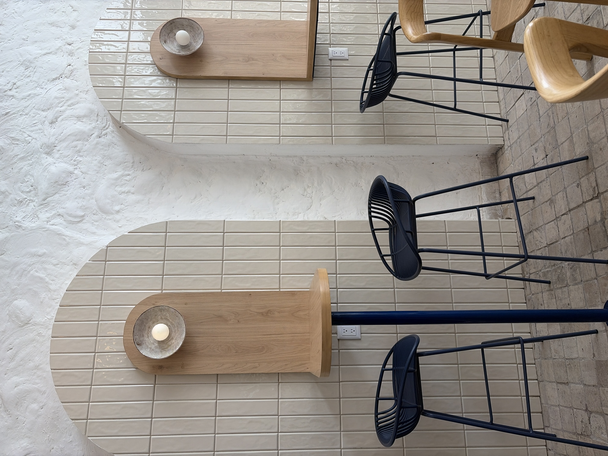 A high-angle, overhead shot captures a seating area with two identical rounded sections. Each section features light beige, rectangular tiles arranged horizontally. Against the textured white wall, each section has a curved edge. On each tiled surface, a light wood-tone shelf is affixed, each holding a shallow, ridged bowl with a light-colored object inside. An electrical outlet is visible on each tiled surface near the shelf. The top seating area has two light wood chairs with black frames. The bottom seating area has two dark blue chairs with black frames, attached to a horizontal dark blue rail. The room appears to be indoors. The location is Unknown, Unknown.