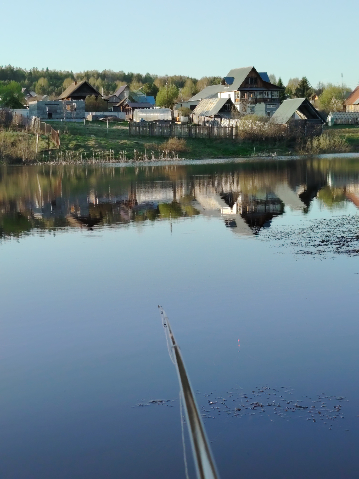 A daytime scene near Марковский, Russia, shows a body of water reflecting a collection of houses and trees. A fishing rod is visible in the foreground, extending into the water, with a red and white bobber floating nearby. Algae or plant matter floats on the surface of the water. The houses appear to be situated on a slight elevation above the water level, with a wooden fence separating the houses from the water's edge.