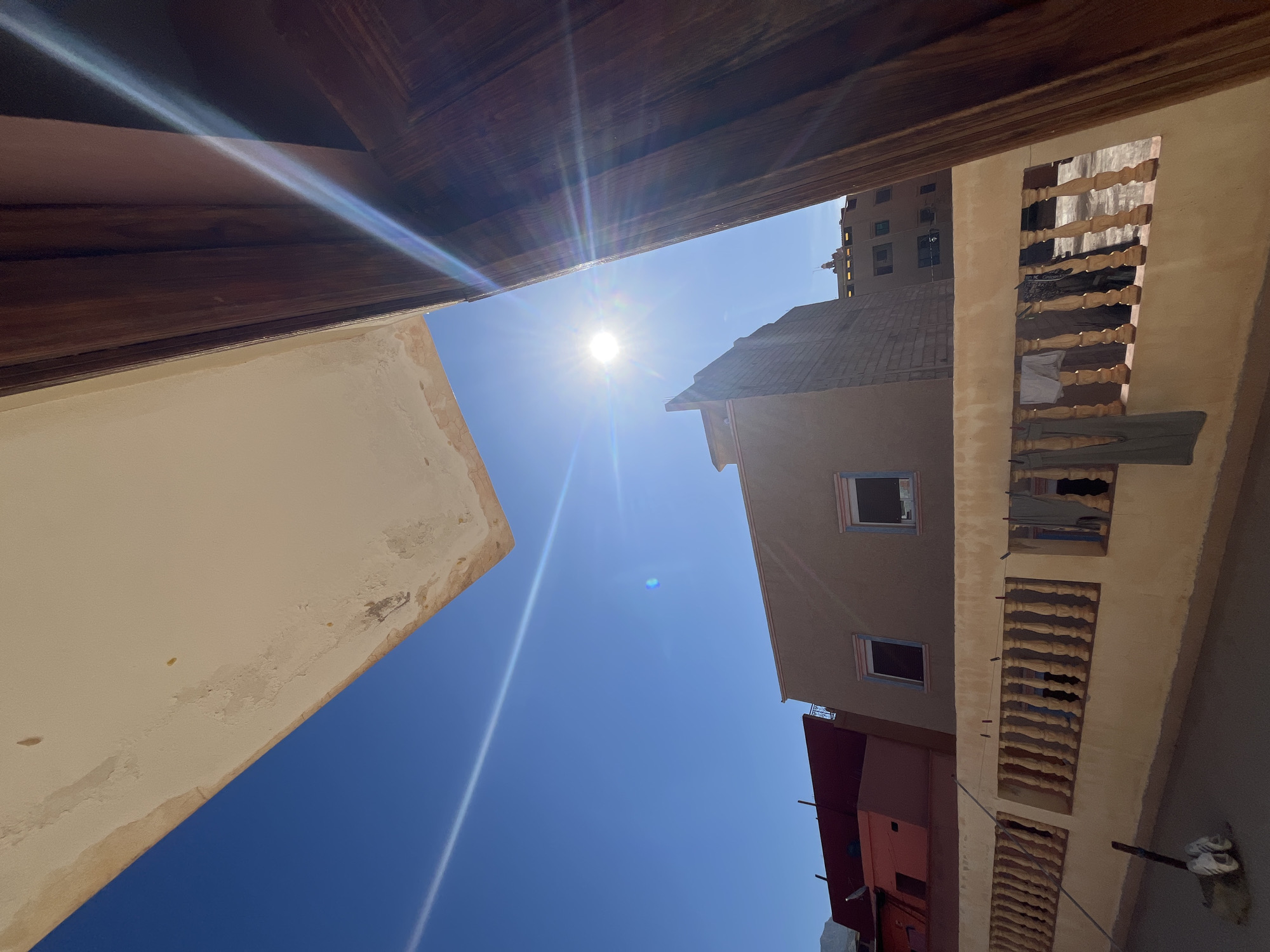 The low-angle shot looks upward from a courtyard, perhaps in the city of Ouaouizert, Morocco. The deep blue sky is pierced by the sun's bright rays. The buildings surround the scene, exhibiting traditional architecture in earth tones, with some decay visible. A building on the right shows clothes hanging out a window. A wooden structure is visible at the top left.