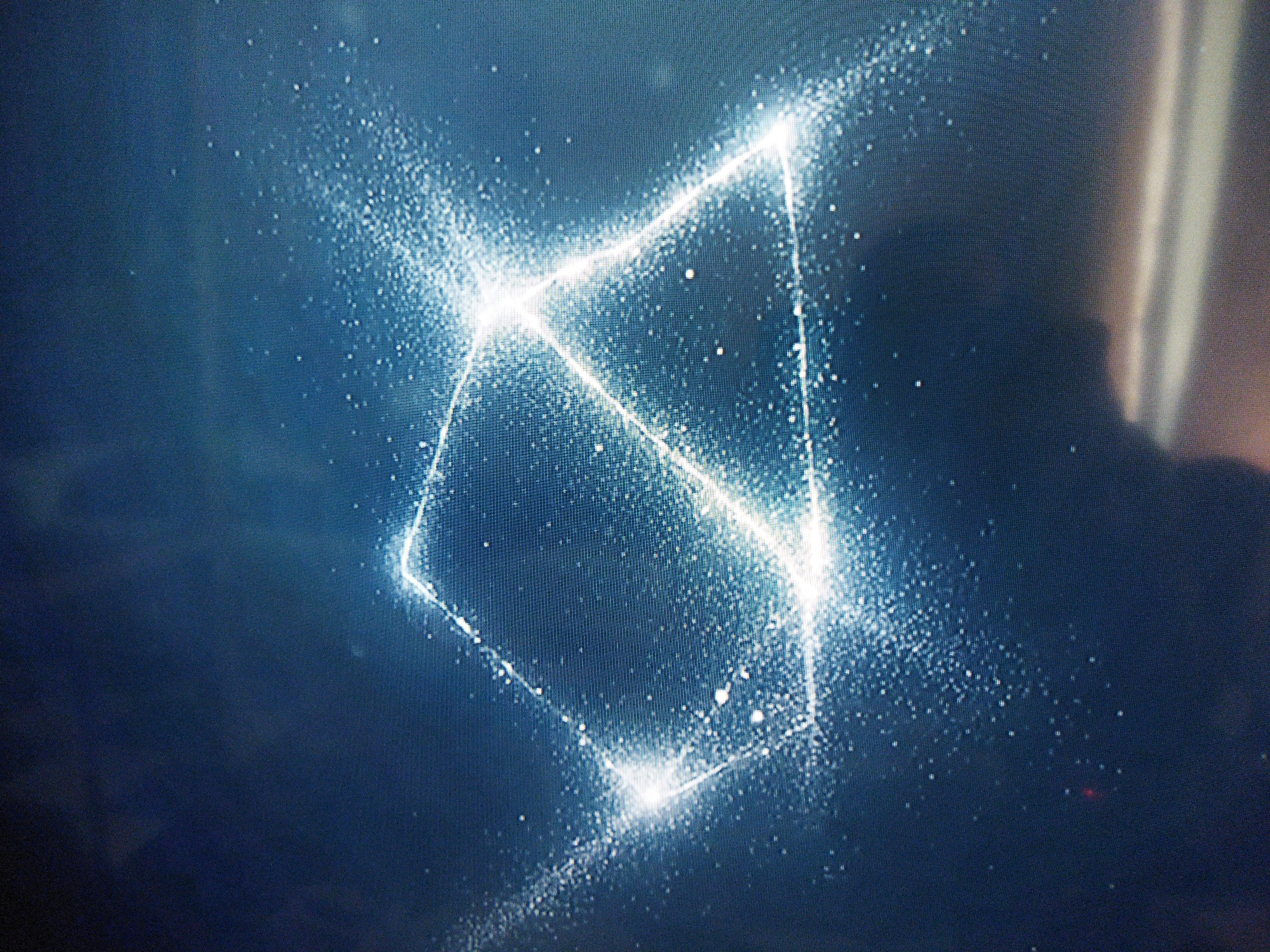 The image shows a glowing geometric shape, possibly a square or diamond, formed from small, bright particles against a dark blue background. The particles are concentrated along the lines of the shape and dissipate outwards, creating a hazy, ethereal effect. Some indistinct shadow forms are visible in the background. The image's perspective is tilted slightly upward, and the focus is soft. There are no discernible details to relate this to Kramatorsk, Ukraine.