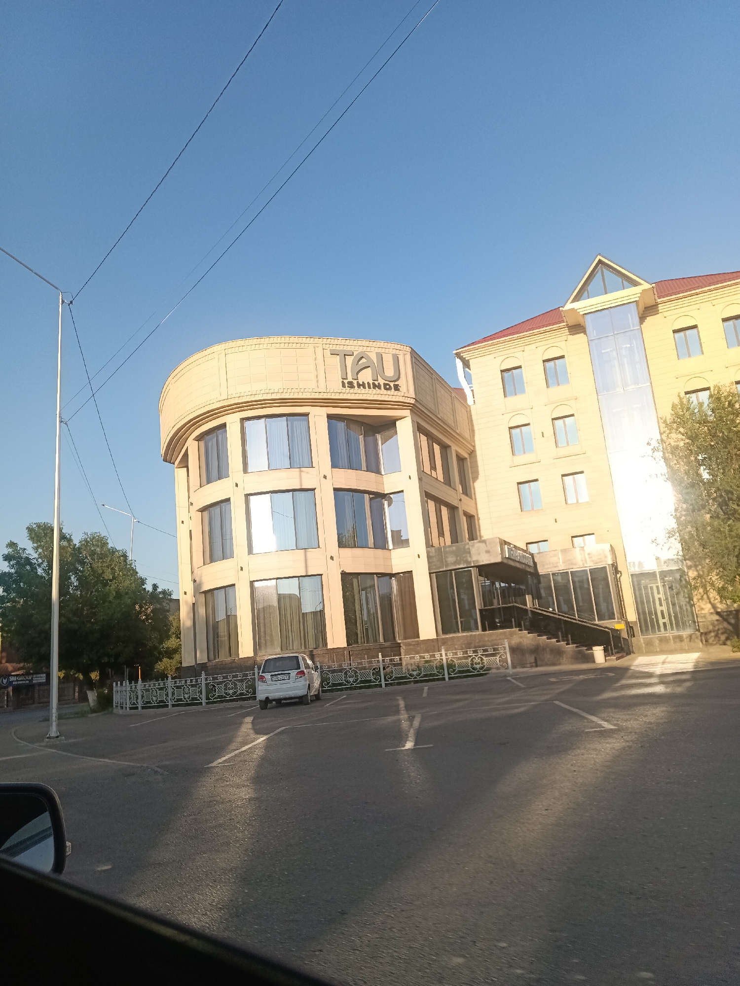 The image shows a modern building with a curved facade on the left and a rectangular section on the right, situated in the city of Shymkent, Kazakhstan. The building appears to be an office or commercial establishment, with large windows and prominent signage that reads "TAU ISHINDE". A white car is parked in front of the building, facing away from the camera. The scene is set against a clear blue sky, with trees and a street visible in the foreground. The lighting suggests it is daytime, possibly late afternoon or early morning, given the long shadows cast on the pavement. The overall impression is one of urban development and a well-maintained commercial area.