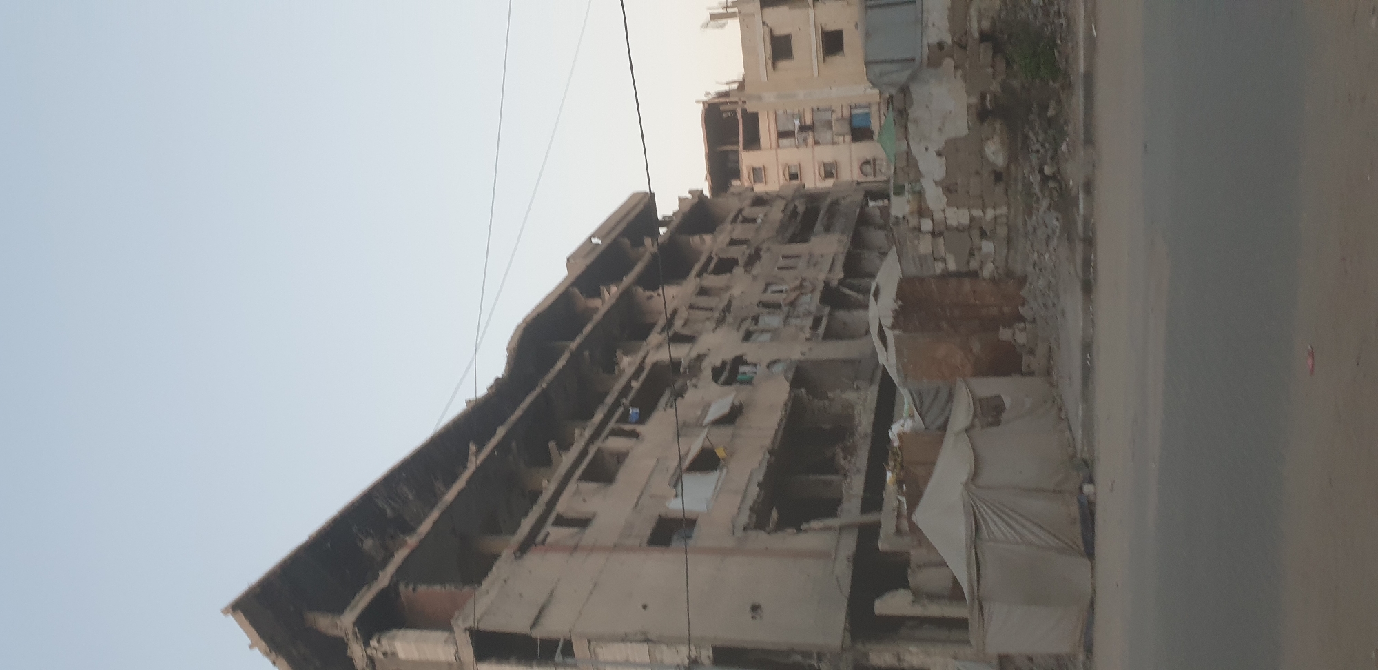 This is a medium-shot, vertical, outdoor photograph of a heavily damaged apartment building in the city of Gaza, Palestinian Territories. The building is multi-storied and appears to be constructed of concrete. Its facade is pockmarked with holes and has sections missing, particularly on the upper floors. The roofline is jagged and broken, with exposed rebar visible. Several windows are present, but many are shattered or empty. Some debris and rubble are visible at the base of the building and in front of it. In the foreground, a street is visible with a curb. There are tents set up in front of the damaged building. To the right of the main damaged building, another multi-storied building is visible, appearing less damaged but still showing signs of disrepair. The sky above is a pale, clear blue. The overall scene conveys a sense of destruction and displacement.