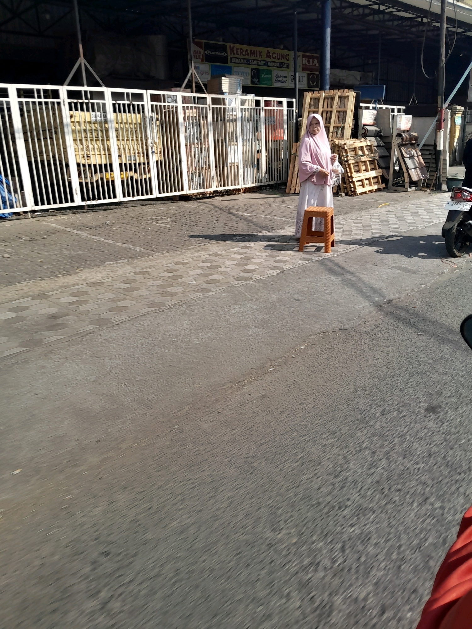 A woman wearing a pink hijab and a light-colored dress stands near a brown plastic stool. She is holding a small white bag. Behind her are stacks of wooden pallets and roofing tiles. A white metal fence is to her left, with a yellow truck visible behind it. The scene appears to be in a commercial or industrial area, possibly a market or a warehouse, in Ngadilangkung, Indonesia, given the presence of signage in Indonesian and the general environment. The ground is paved with interlocking concrete blocks and asphalt. A motorcycle is parked to the right of the woman.