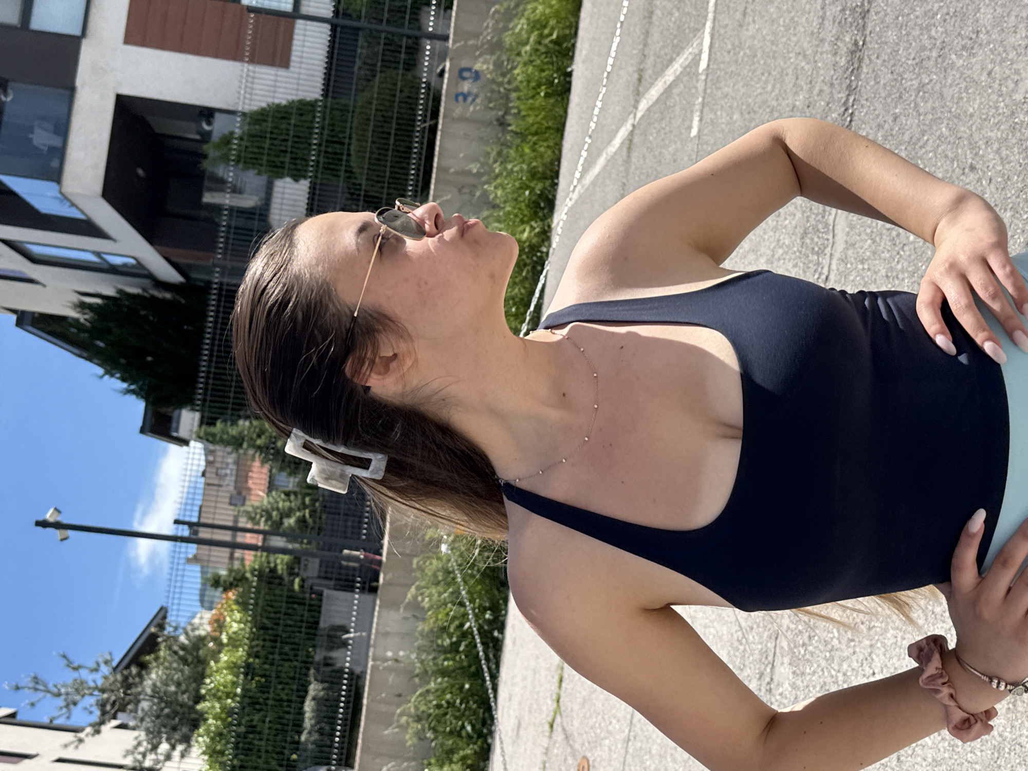 A full shot shows a young woman outdoors in Sofia, Bulgaria. She is fair-skinned, with sunglasses and a hair clip. She wears a black tank top and a light-colored bottom. She also wears a necklace, bracelets, and hair scrunchies. She poses with her hands on her hips, looking upwards. The background features modern apartment buildings, a wire fence, trees, and a paved area. The sky is blue, suggesting a sunny day.