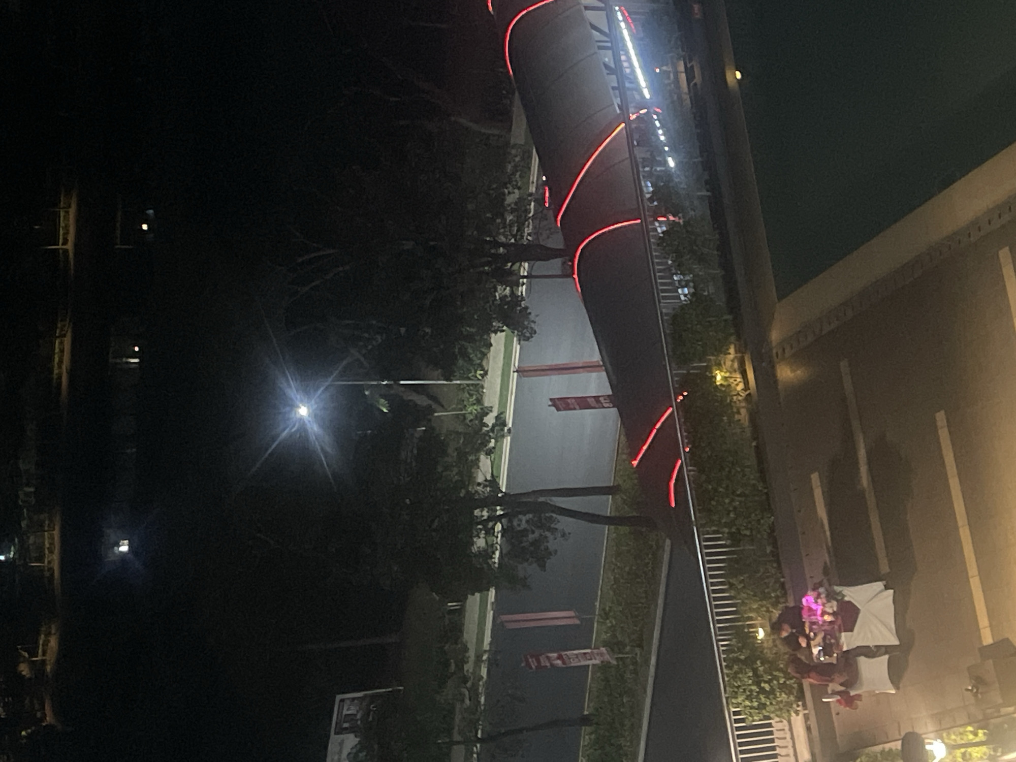 An overhead shot captures a romantic outdoor dinner setup at night in the city of Bekasi, Indonesia. Two people are seated at a table adorned with a red tablecloth, a bouquet of pink flowers, and what appears to be a small umbrella or decoration. The scene is illuminated by ambient light, including a bright streetlamp visible in the background. In the middle ground, a walkway or bridge with red neon accents curves across the frame, separating the dining area from a street lined with trees and signage. The foreground features a portion of a swimming pool, its dark water reflecting minimal light. The overall impression is one of an intimate dining experience set against a backdrop of urban infrastructure and nighttime ambiance.