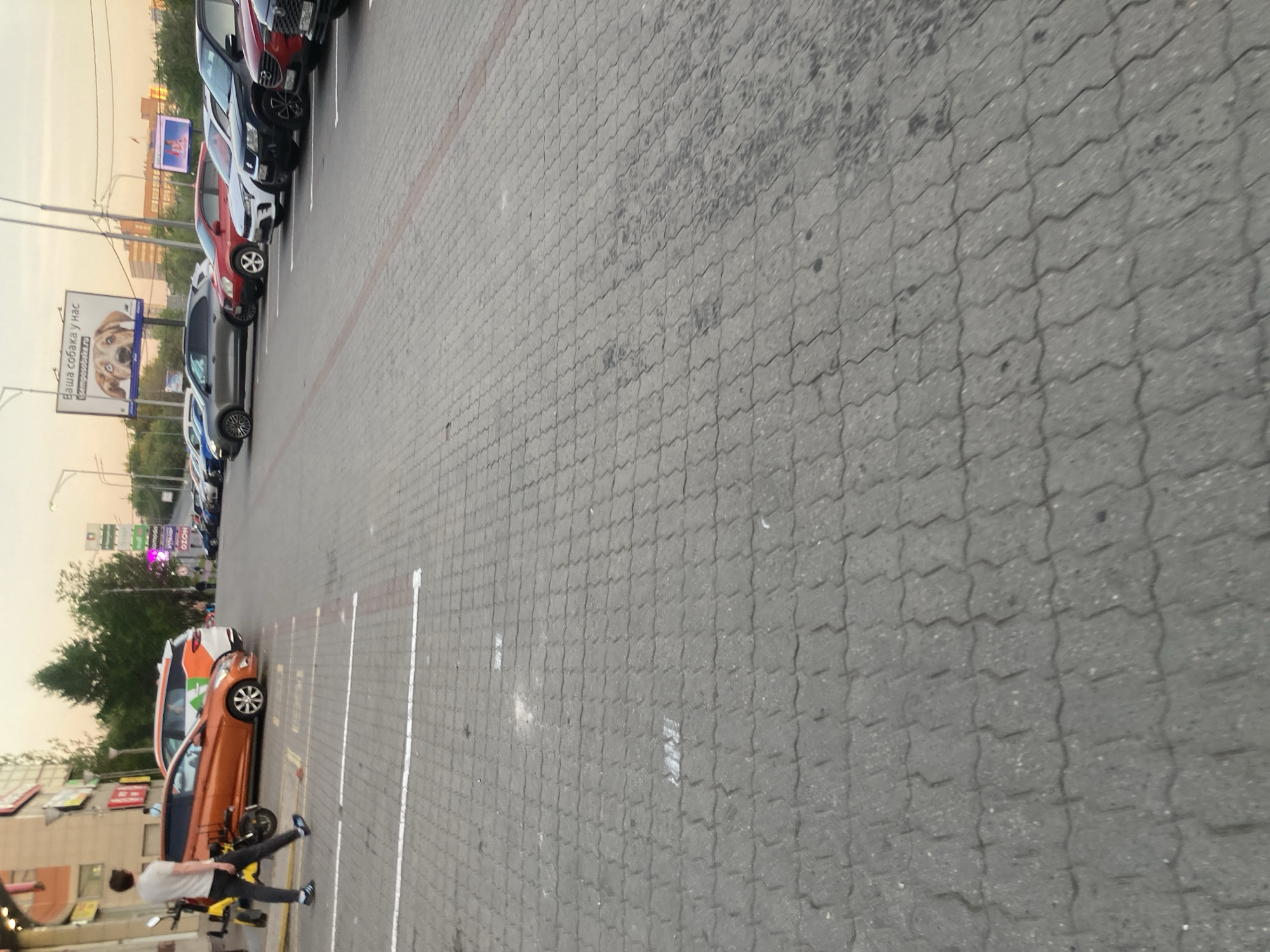 Here is a description of the image.
The image shows an outdoor parking lot paved with gray interlocking bricks. The parking lot appears to be in Moscow, Russia, based on the billboard with Cyrillic text. Rows of parked vehicles are visible, including cars of various colors like red, white, gray, and orange. A person wearing a light-colored shirt and dark pants is seen near an orange car, next to a scooter. Street lights and trees are visible in the background. Buildings are also present across the street. The sky is overcast.