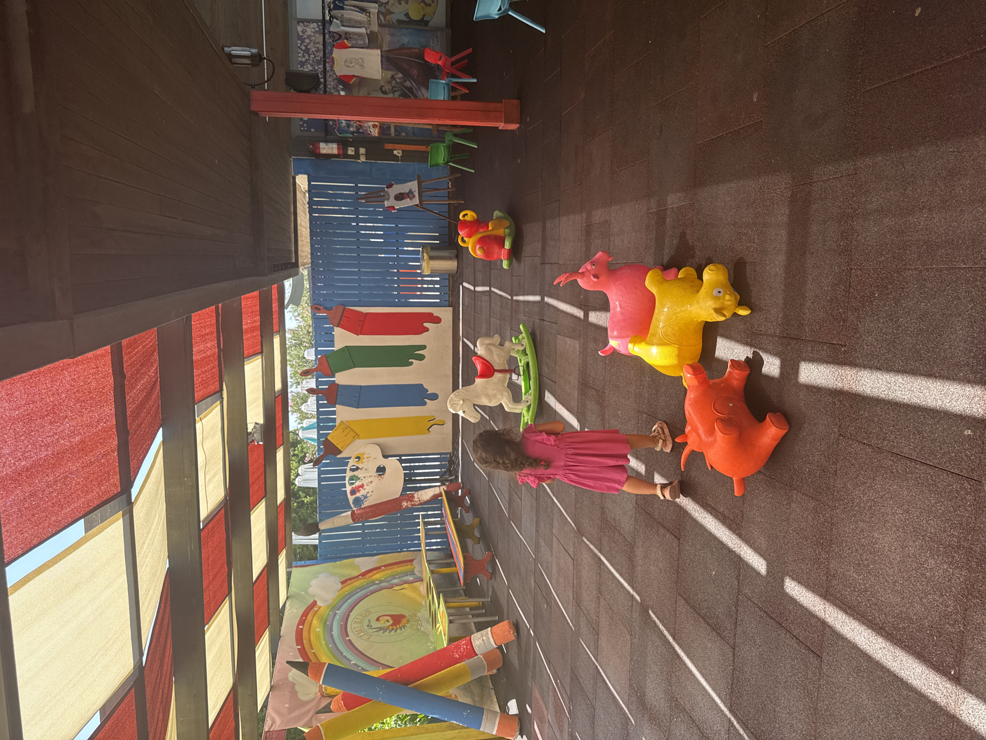 An overhead canopy with alternating red and beige fabric sections covers an outdoor play area in Aksu, Turkey. A young girl in a pink dress stands facing away from the camera, looking towards a blue wooden fence decorated with painted oversized art supplies like pencils, paintbrushes, and a palette. The ground is a dark rubberized surface with bright white lines creating a striped pattern. In the foreground, three colorful inflatable animal toys are scattered on the ground: an orange pig-like creature, a pink goat-like creature, and a yellow dog-like creature. Further back, a white rocking horse with a red saddle sits on a green base. Various tables and benches are positioned in front of the blue fence. Other playground equipment, including red and green chairs and a red fire extinguisher, are visible in the background.